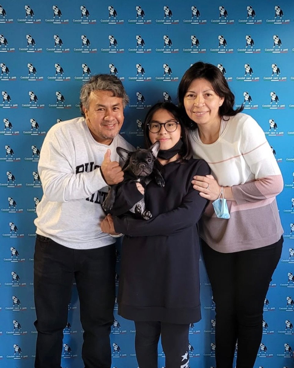 Una famiglia con due adulti, una ragazza con occhiali e un cagnolino, posano sorridenti davanti a uno sfondo con un logo di un cane bianco e nero e la scritta 'Cuccechi cani e gatti'.