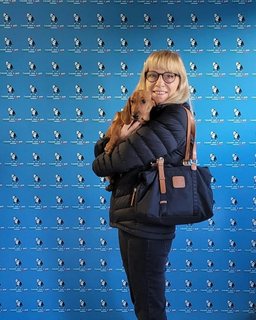 Una donna con occhiali tiene un cucciolo di cane marrone mentre sorride davanti a uno sfondo blu con loghi di un panda che corre, e il testo 'CaniCani e Gatti' scritto più volte.