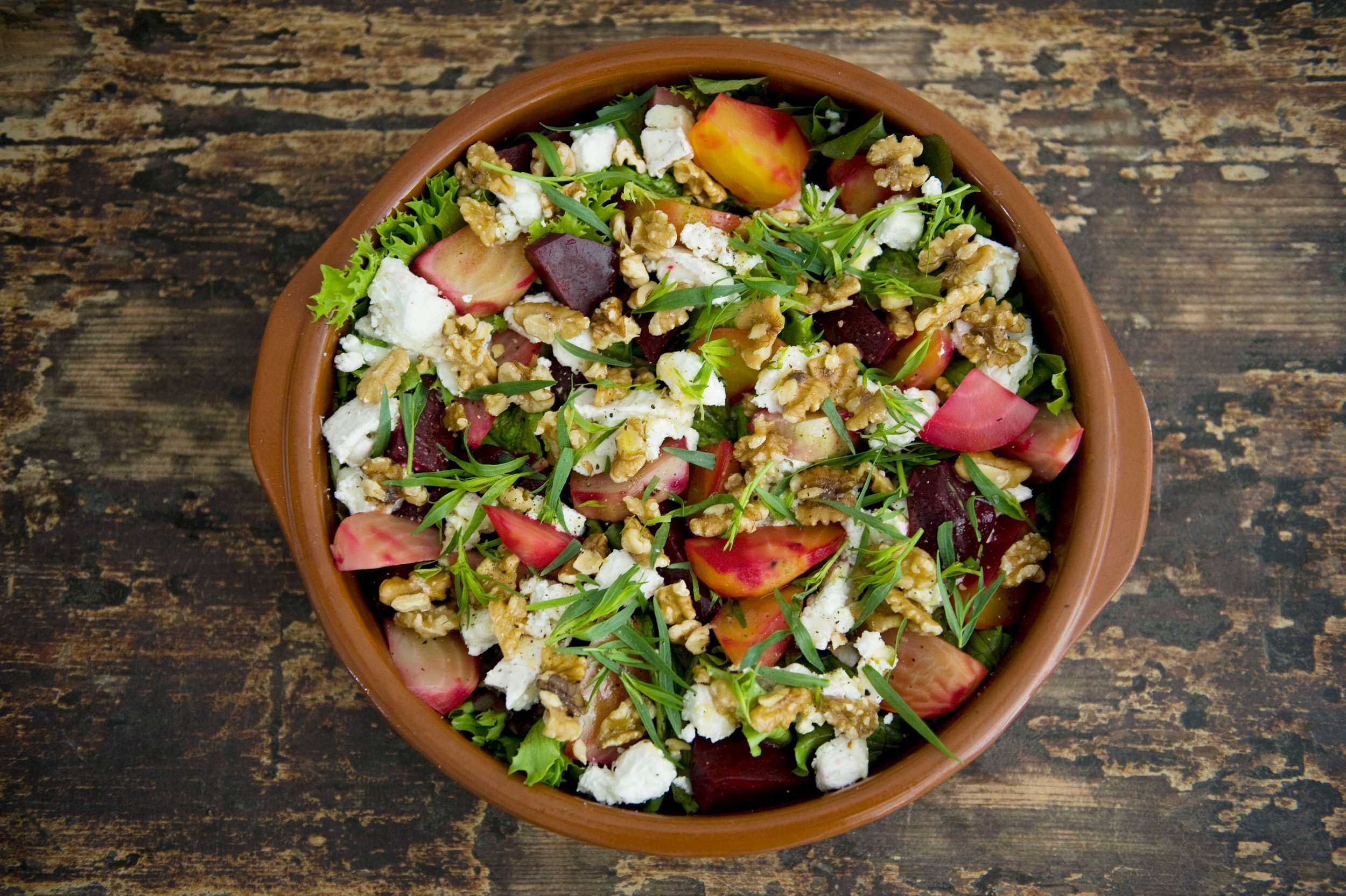 A bowl of mixed salad with sliced radishes, beets, crumbled cheese, walnuts, and greens on a wooden surface.