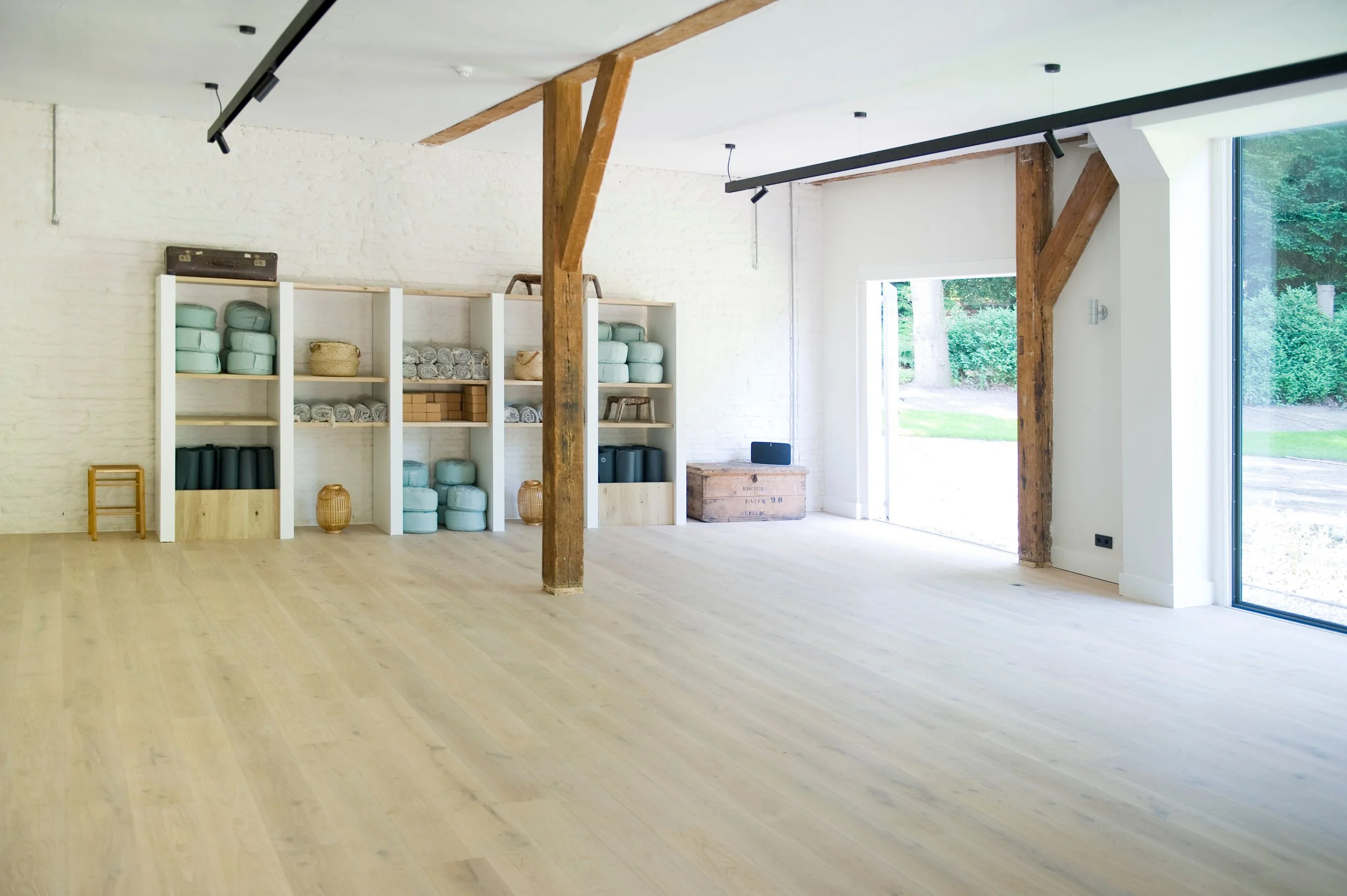 An empty room with light wooden floors and white walls, featuring open shelves stocked with exercise mats, towels, and baskets, large windows, and wooden beams.