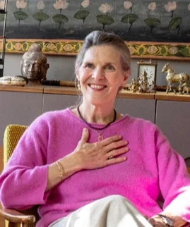 A smiling woman with short gray hair, wearing a pink sweater and white pants, sitting indoors with her hand on her chest.