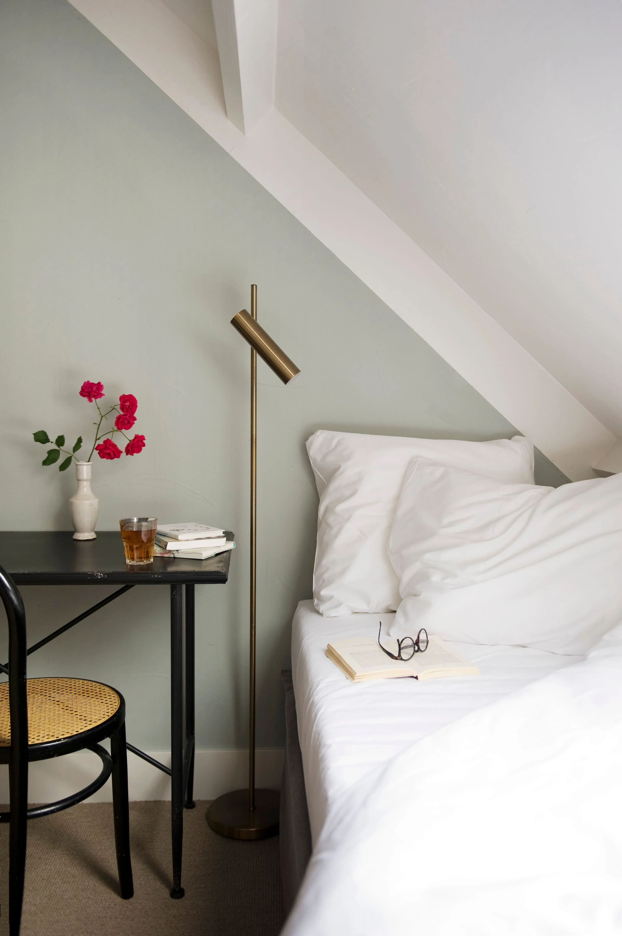 Retreat Christy Linle |cozy bedroom corner with a white bed, open book and glasses on it, next to a side table with a vase of pink flowers, a glass of tea, and books, illuminated by a gold floor lamp, against a light green wall with a sloped ceiling.