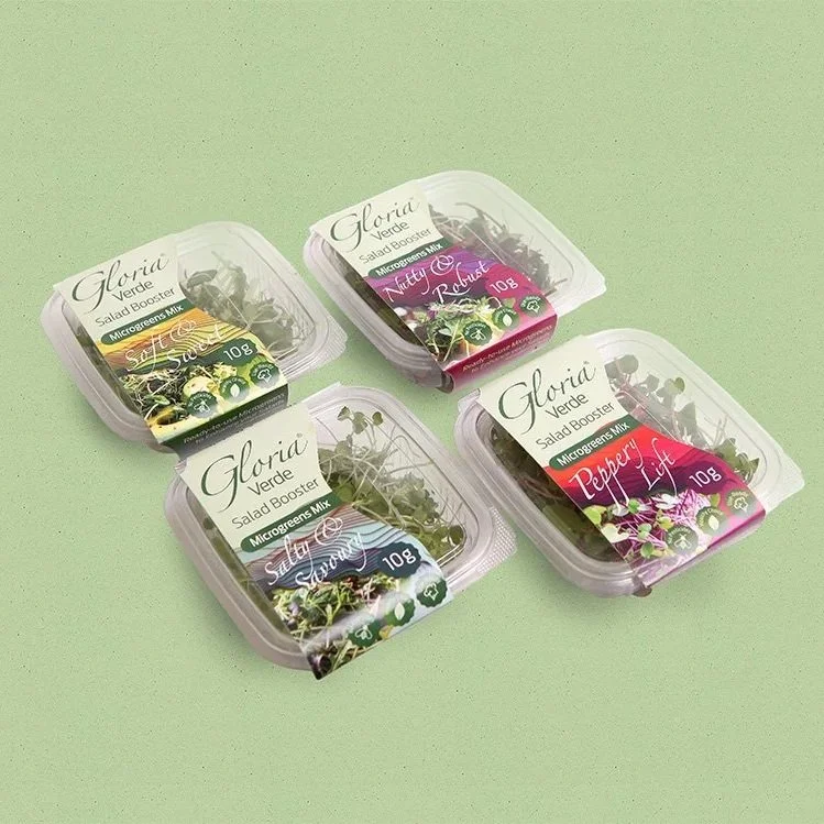 Four small clear plastic containers of salad greens with labels indicating different mixes, placed on a light green surface.
