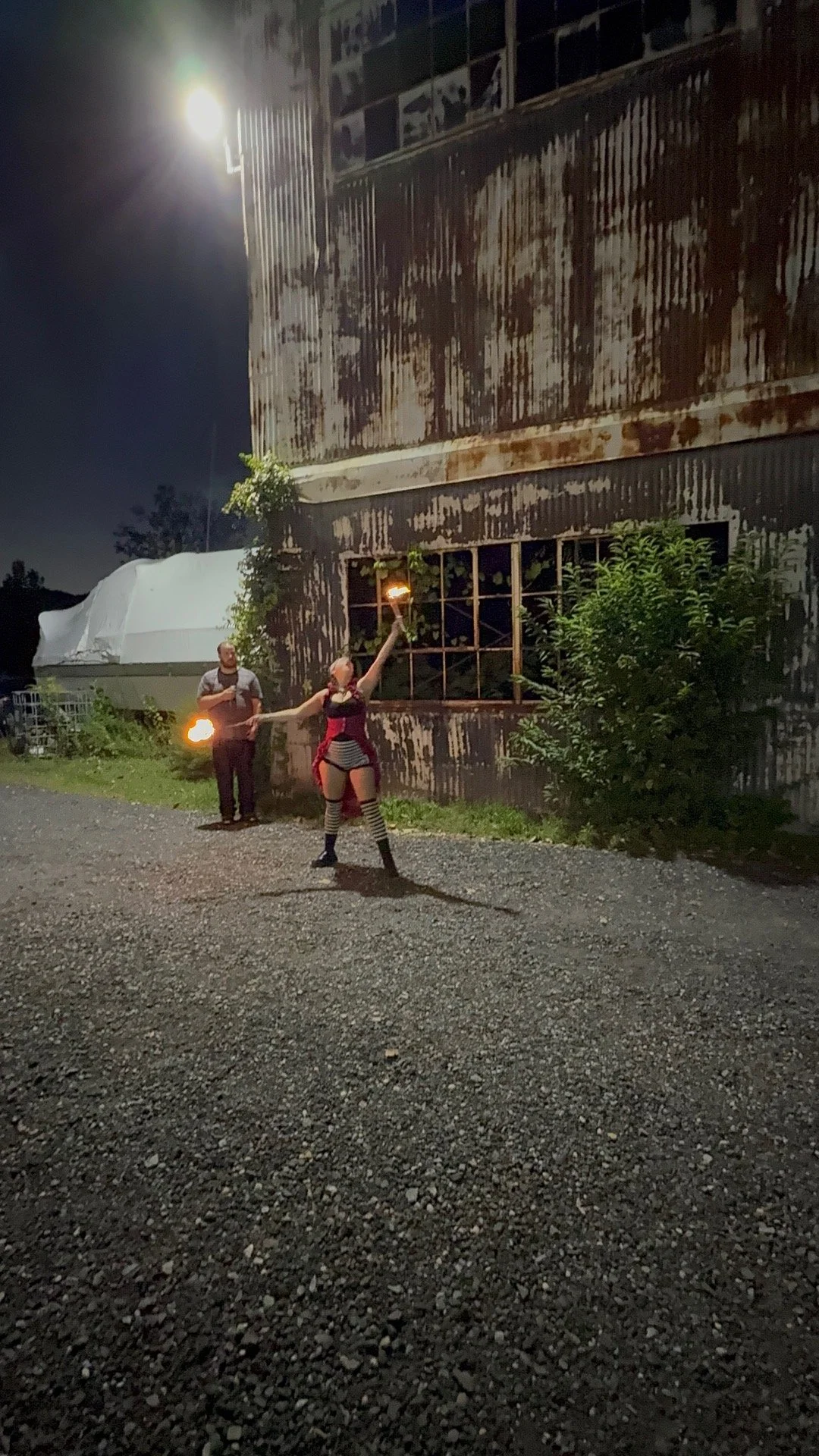A street performer juggling fire outdoors at night, with two men watching in front of a weathered, rusty building and a large white tent in the background.