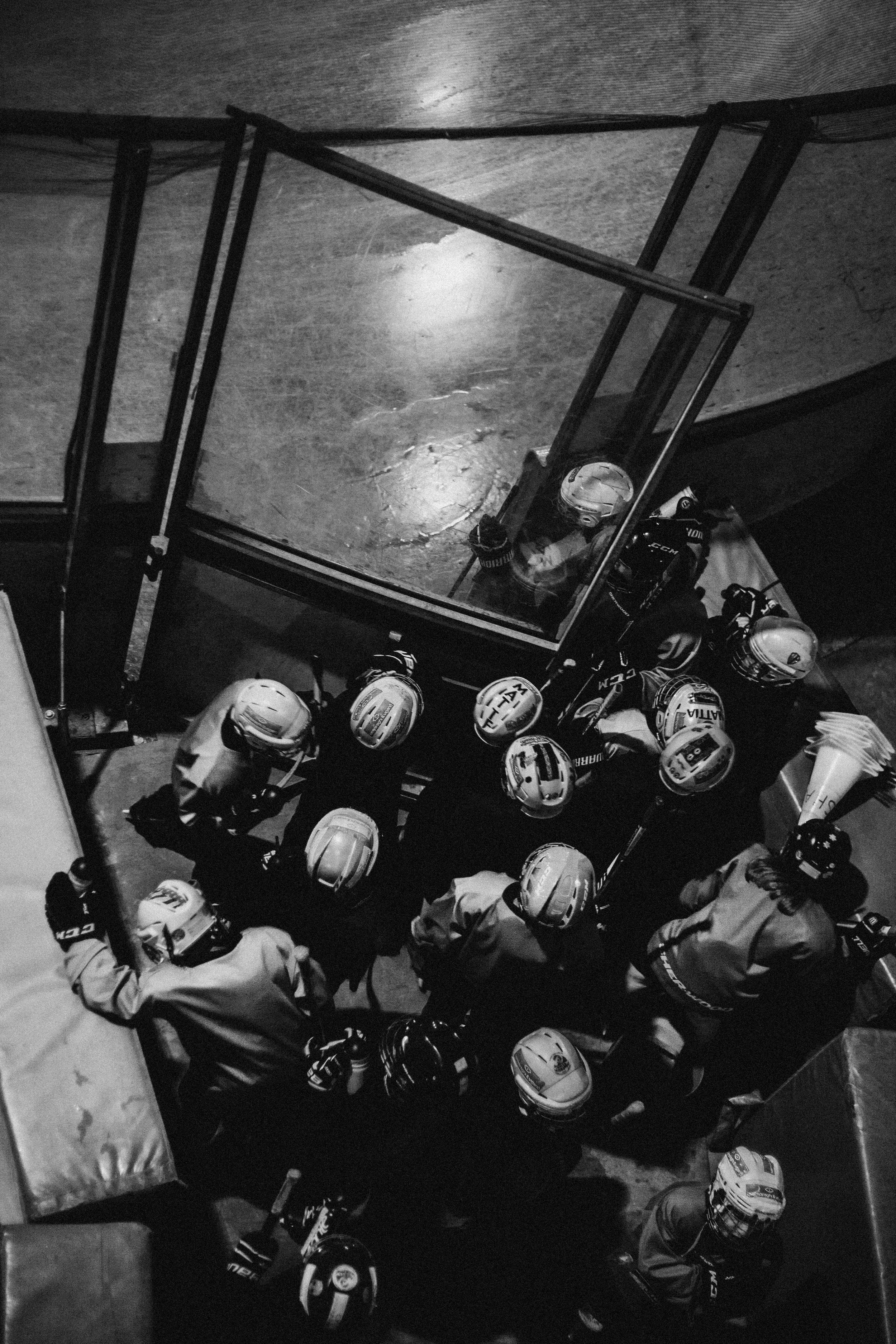 A black-and-white photo taken from above of hockey players huddled together before a game or practice, surrounded by helmets and gear, with a coach or official nearby.