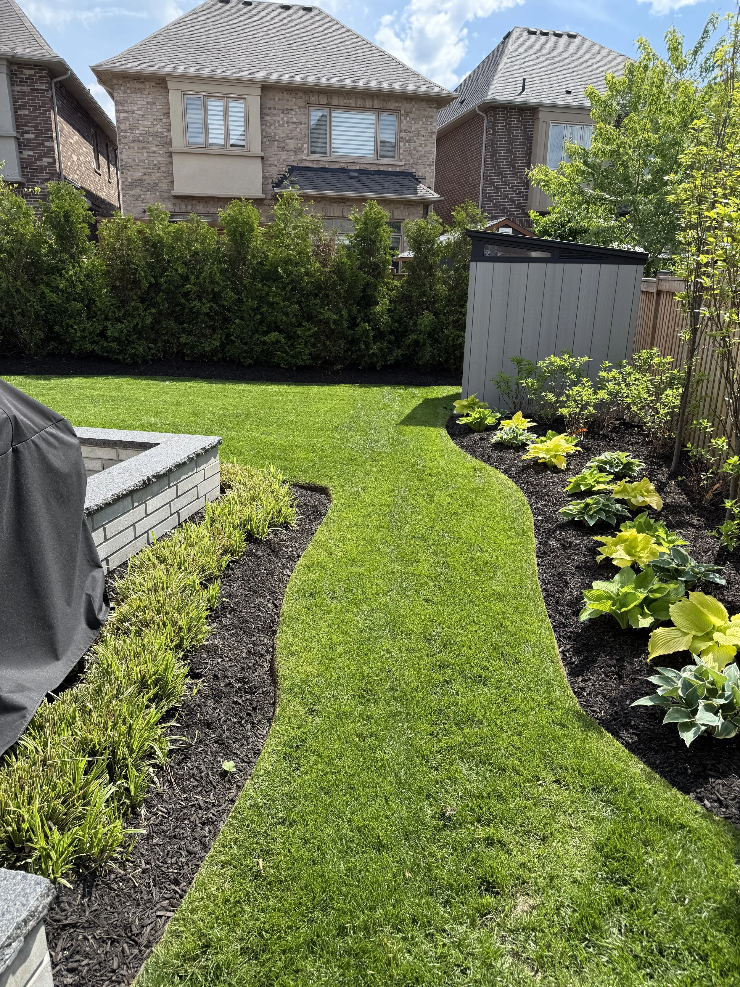 A well-maintained backyard with a curved grass pathway, garden beds with various plants and shrubs, a small garden shed, and neighboring houses in the background under a partly cloudy sky.