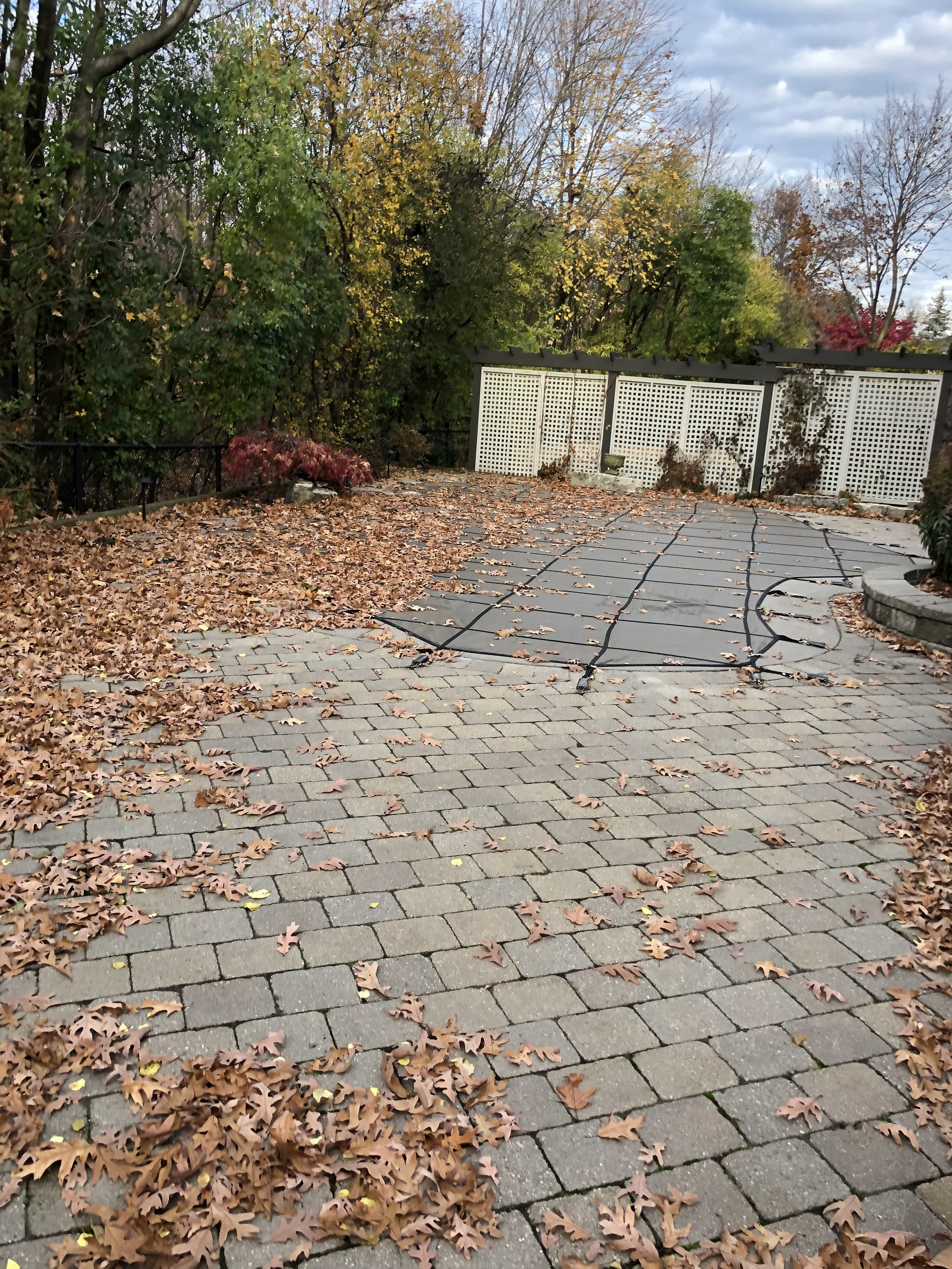 Uncovered patio with brick pavers and fallen autumn leaves, surrounded by a low stone wall on the right, black metal fence on the left, with covered hot tub or pool in the background, and trees with fall foliage and cloudy sky overhead.
