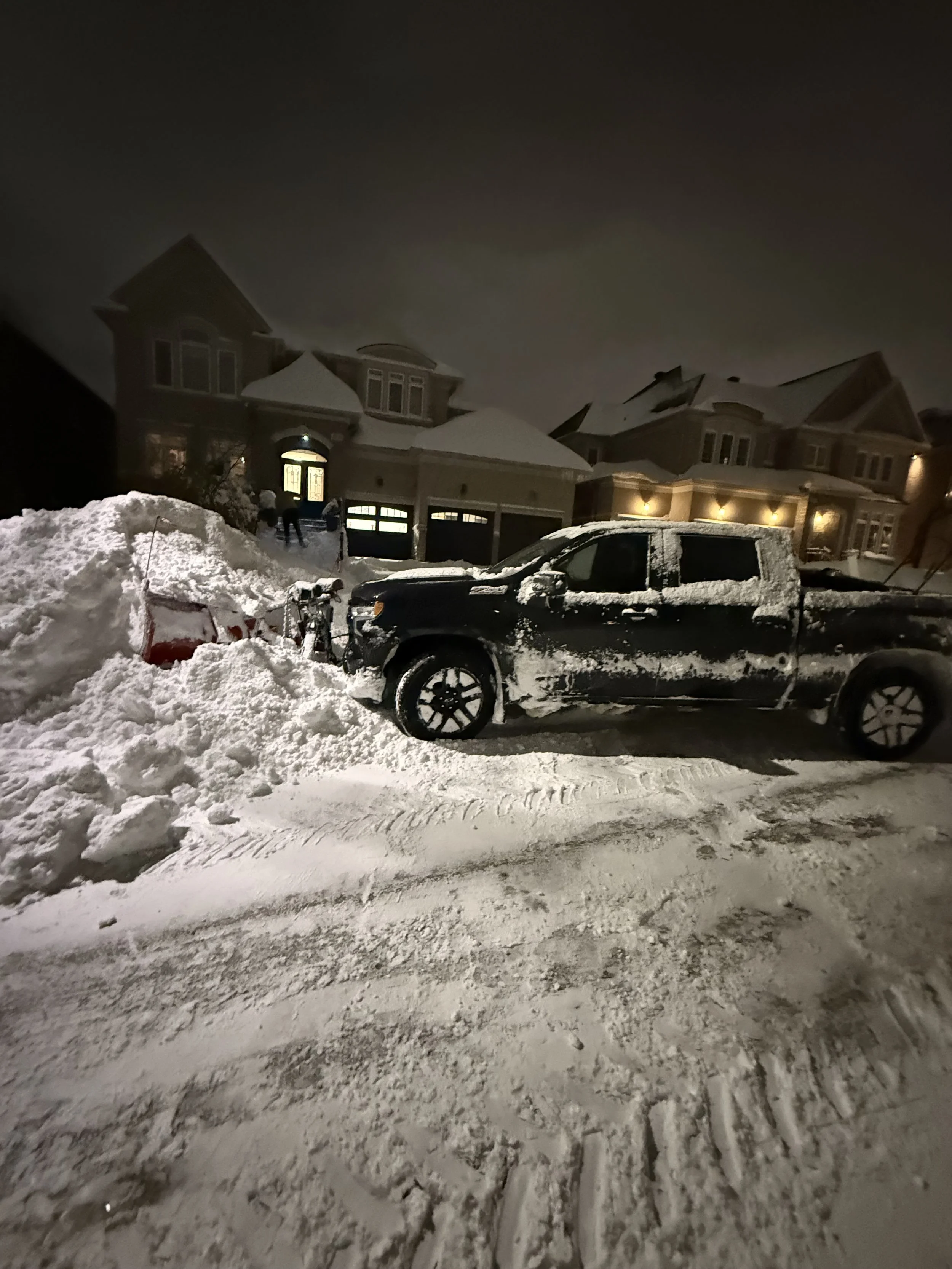 A black pickup truck parked in a snowy driveway near a residential house at night, with snow piled up on the sides and snow covering parts of the vehicle.