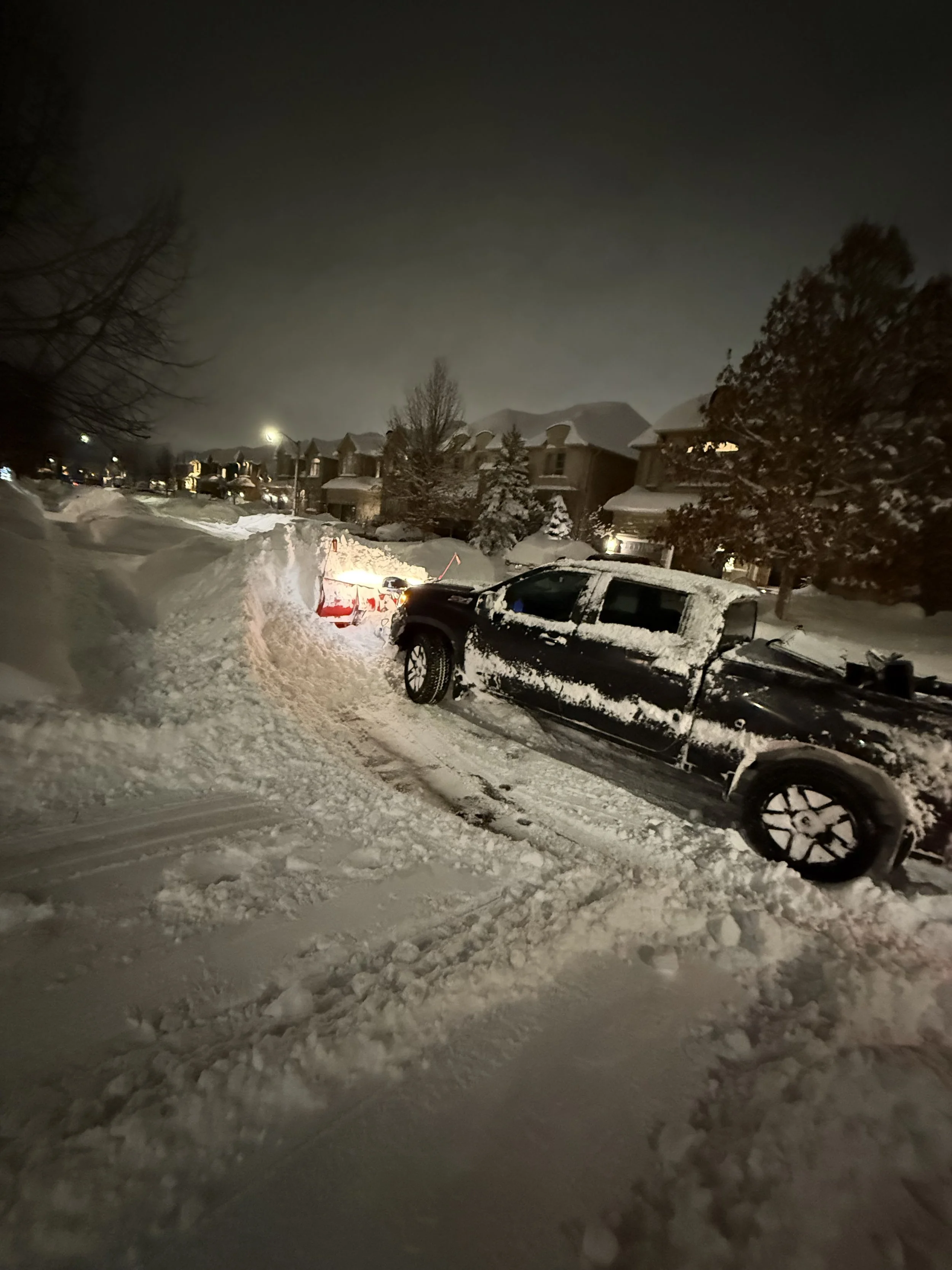 A snow-covered residential street at night with a black pickup truck partially buried in snow and a red snowplow clearing the road.
