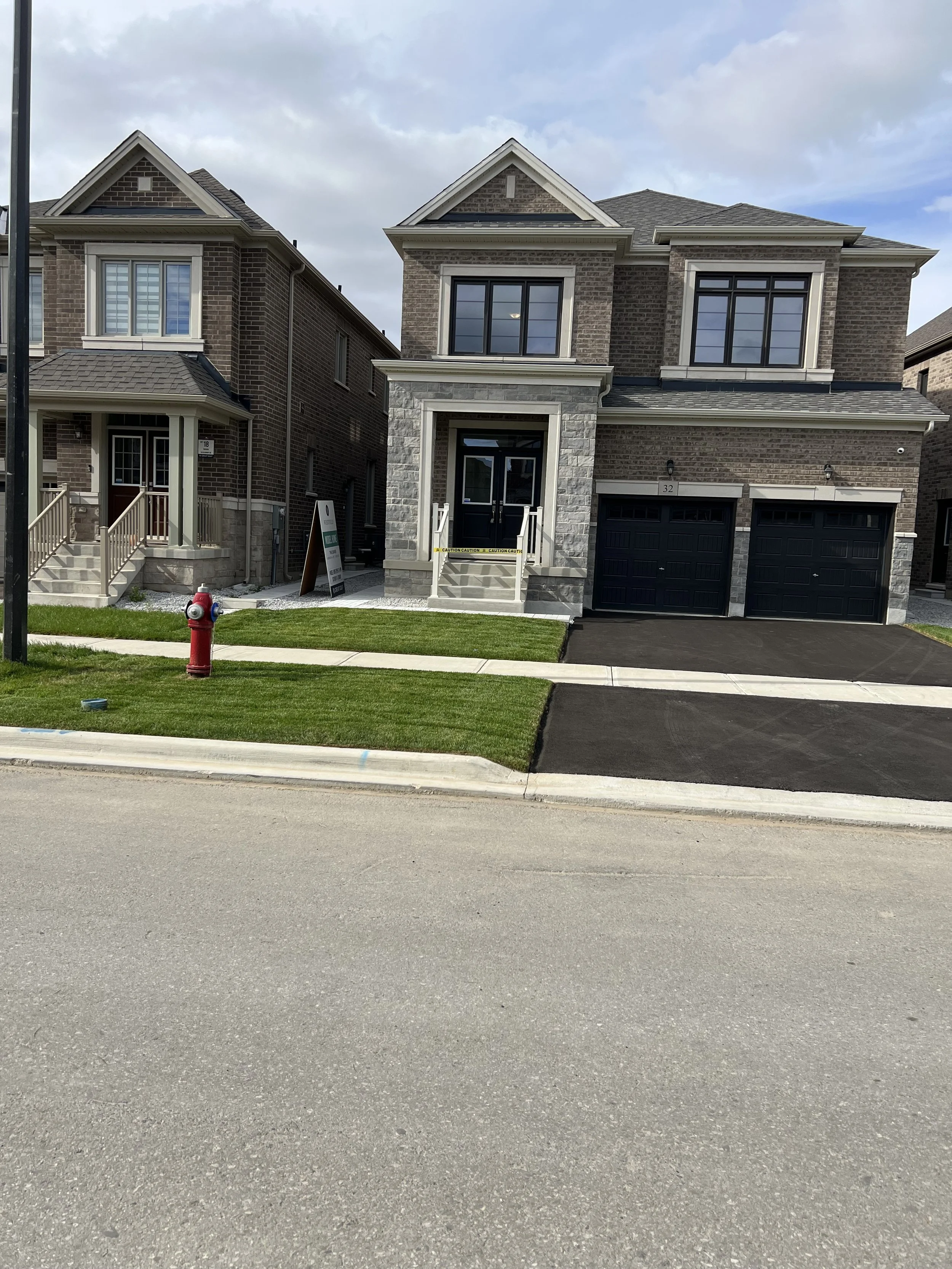 A newly built two-story brick house with a double garage, front steps, and a small front yard with grass, sitting on a residential street under a cloudy sky.