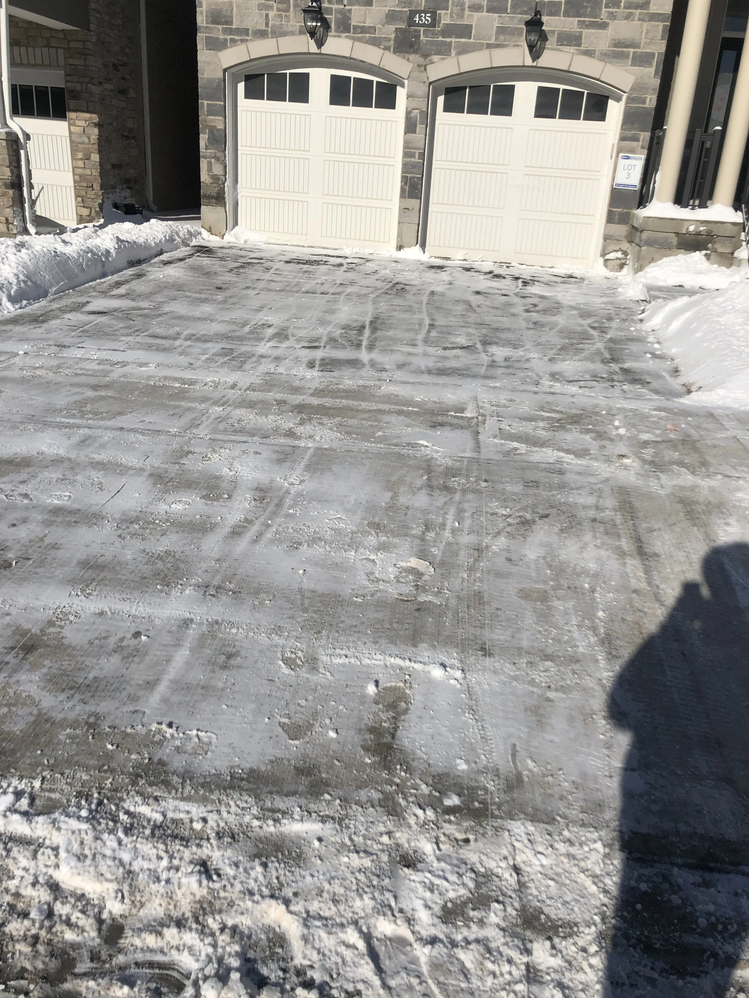 Snow-covered driveway leading to a two-car garage with white doors and stone exterior in a residential area during winter.