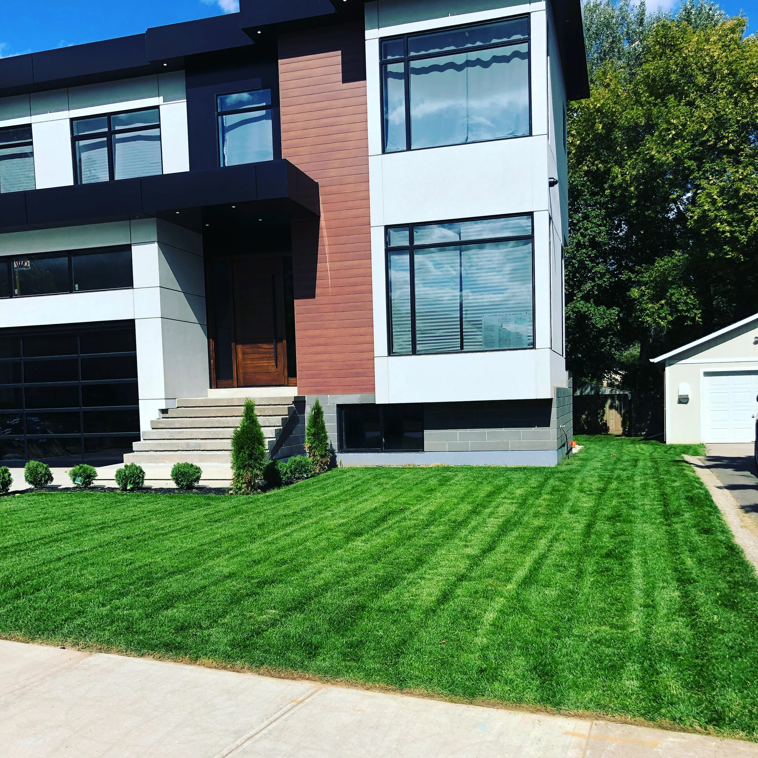 Modern multi-story house with a well-maintained front lawn, large windows, and stairs leading to the front door, with a small white garage in the background.