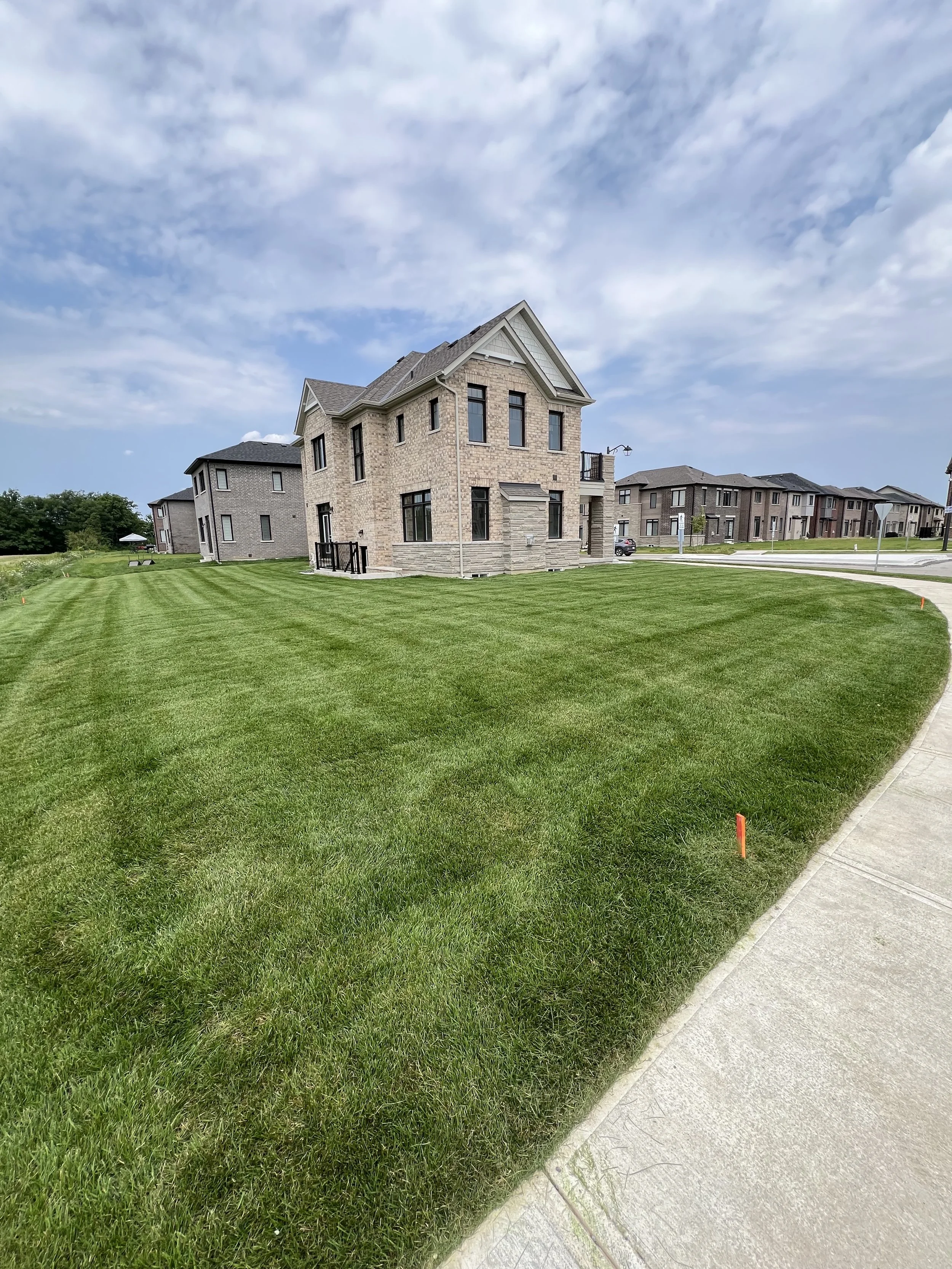 A modern two-story house with brick exterior, multiple large windows, and a small deck, situated on a well-maintained lawn with neighboring houses visible in the background under a partly cloudy sky.