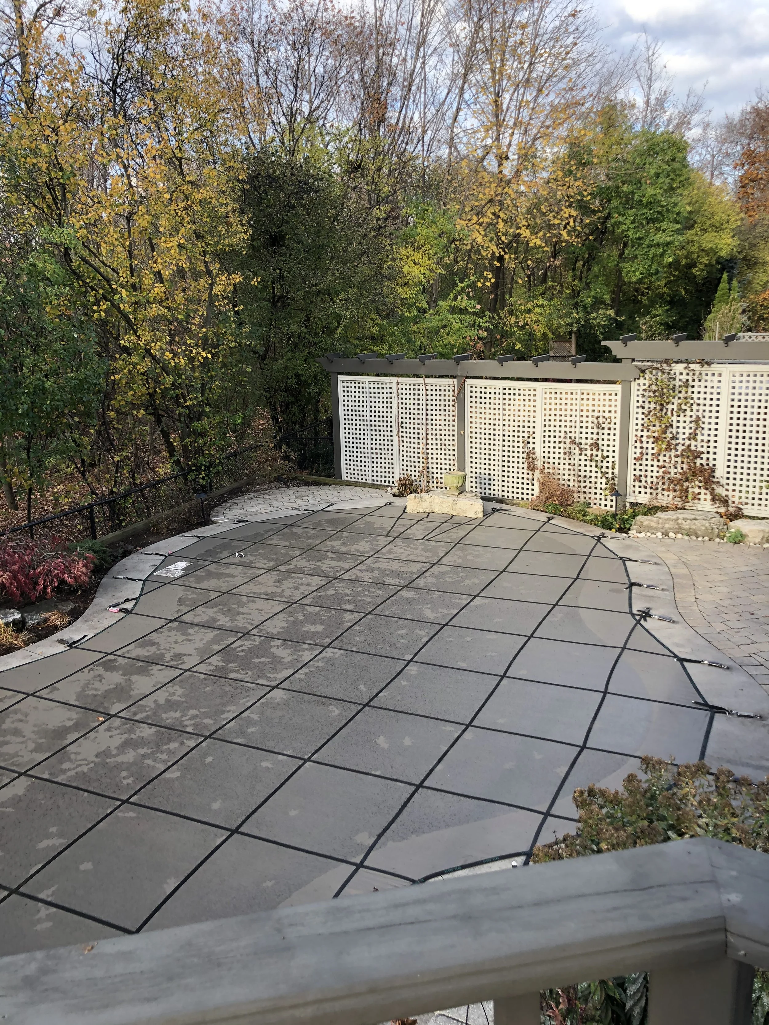 An outdoor patio area with a concrete surface, covered by a black mesh solar pool cover, bordered by a white lattice fence, with trees and foliage in the background during fall.