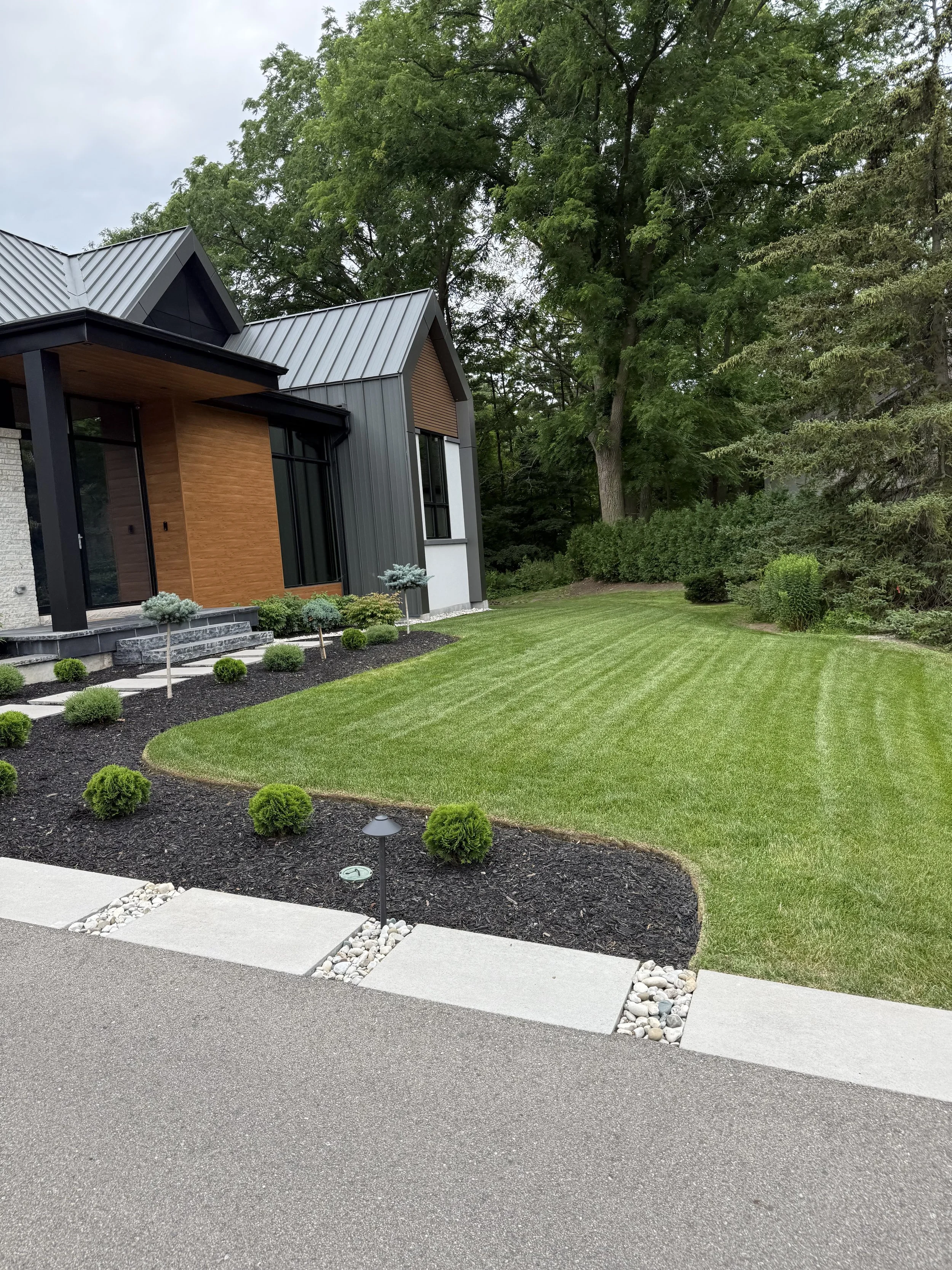 Modern house with metal roof, wood and stone exterior, well-maintained lawn, small shrubs, and trees in the background.