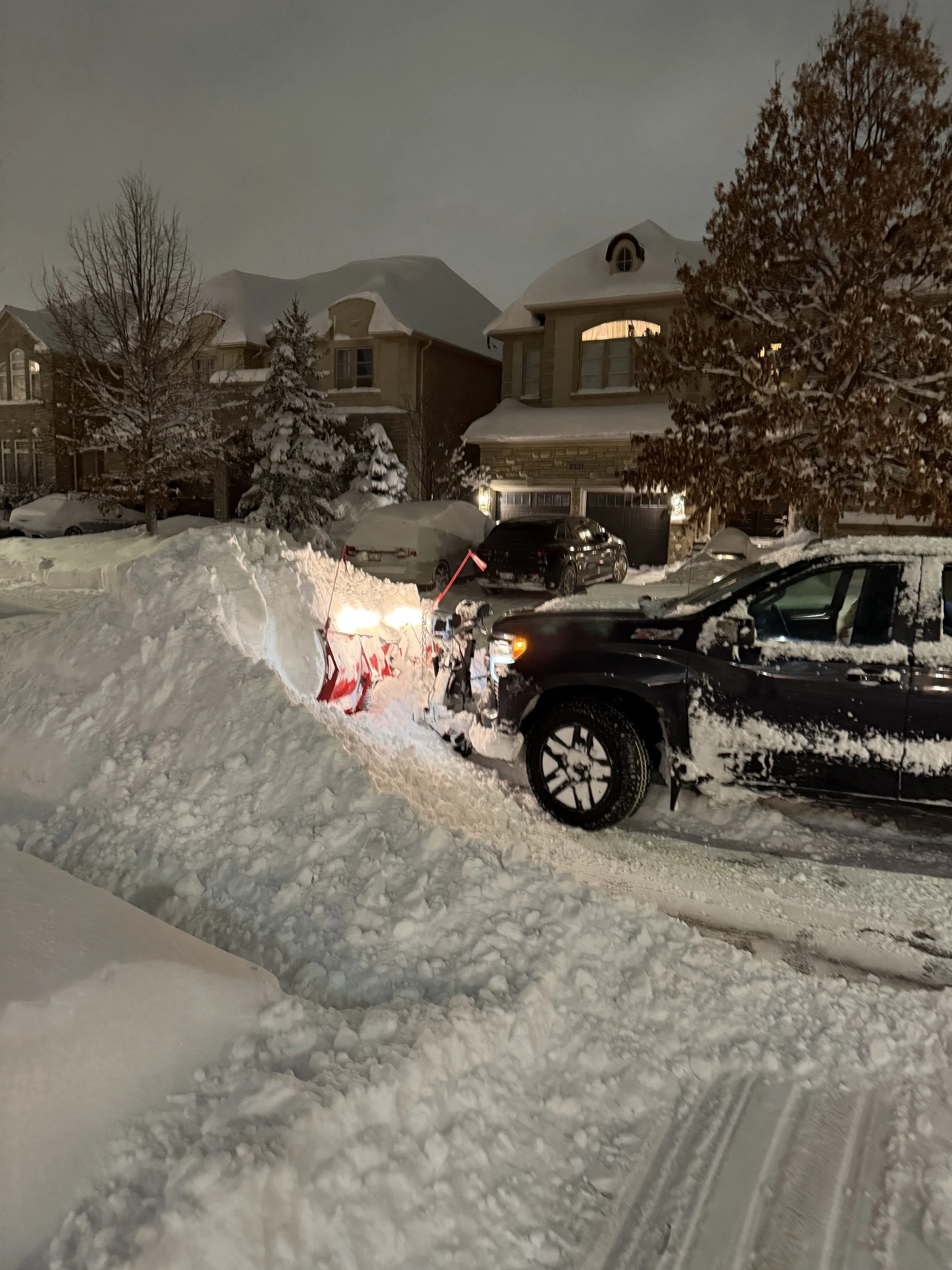A snowplow clearing snow from a residential street at night, with snow piled up around it and snow-covered houses and trees in the background.
