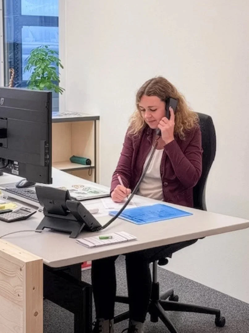 Frau arbeitet im Büro, spricht am Telefon, schreibt Notizen, sitzt am Schreibtisch mit Computer, Telefondisplay und Unterlagen.