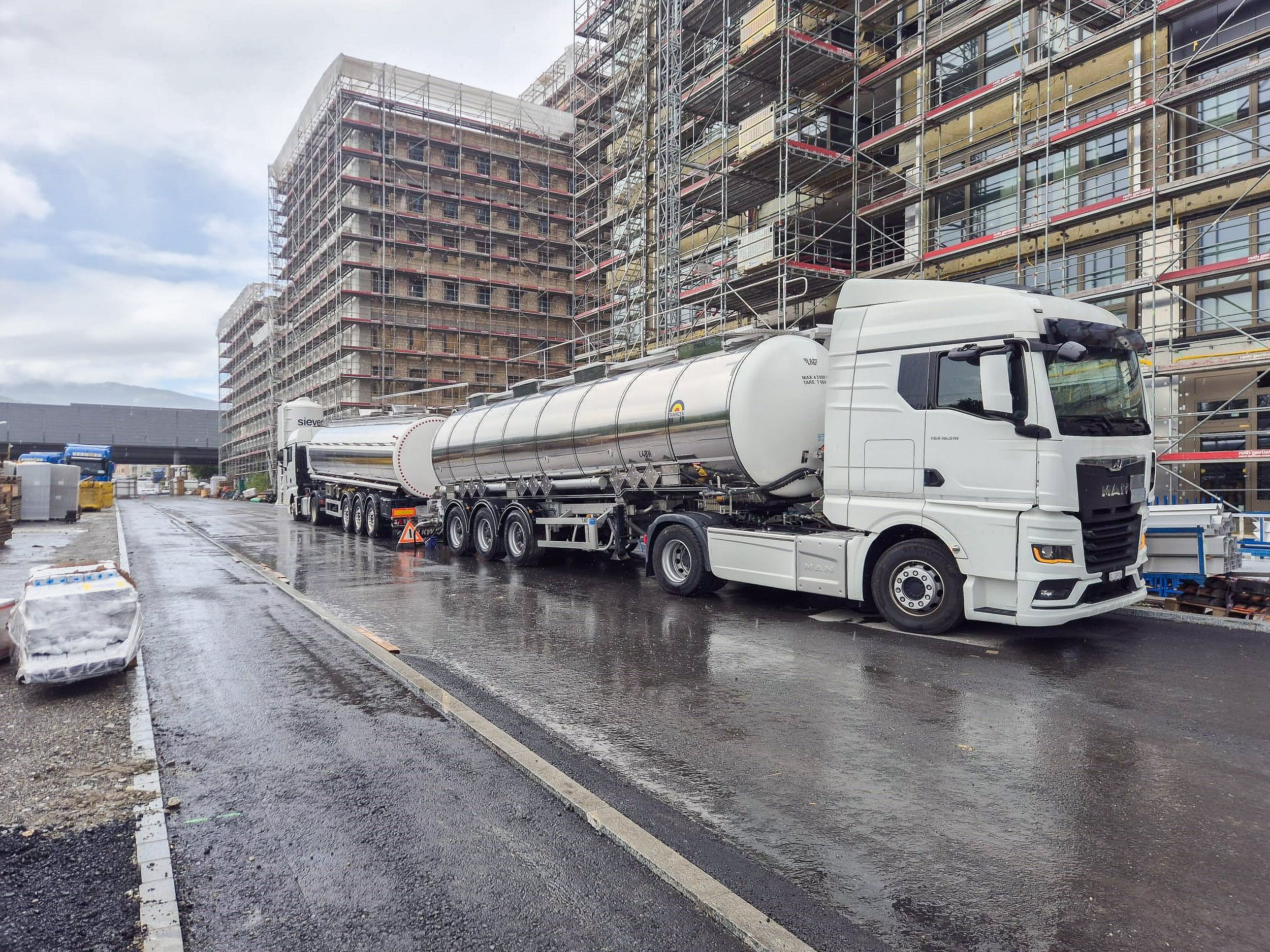 Ein weisser Tanklastwagen steht vor einem Baugerüst an einem Baustellengelände, die Strasse ist nass vom Regen, und es sind Baumaterialien und Verpackungen auf dem Boden zu sehen.
