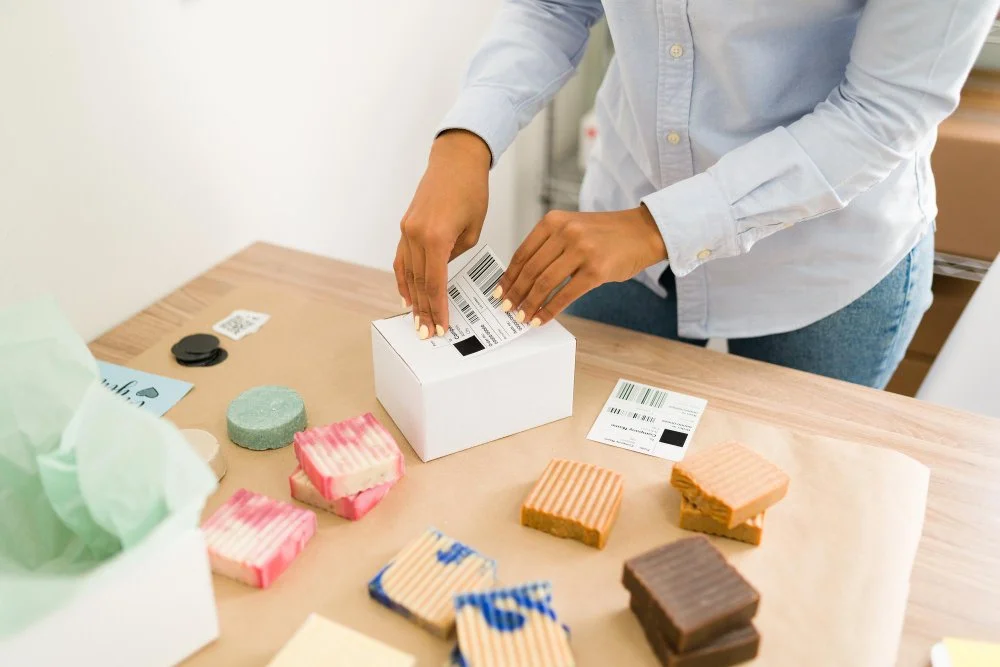 Person packing a box with mailing labels in a workspace with various soap bars and packaging supplies on a wooden table.
