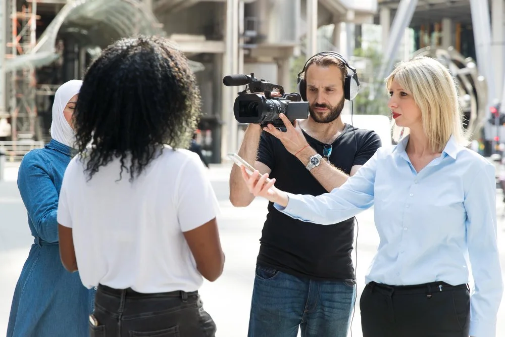 A diverse group of four women and a man are on a film set outdoors during the daytime. The man is operating a professional video camera, wearing headphones, and is focused on filming. The women are engaged in a discussion, with one woman holding a smartphone, another standing with her arms crossed, and the others observing.