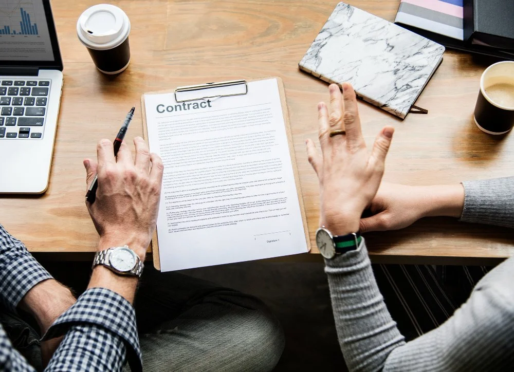 Two people reviewing a contract on a clipboard at a wooden desk, with laptops, notebooks, coffee cups, and a pen.