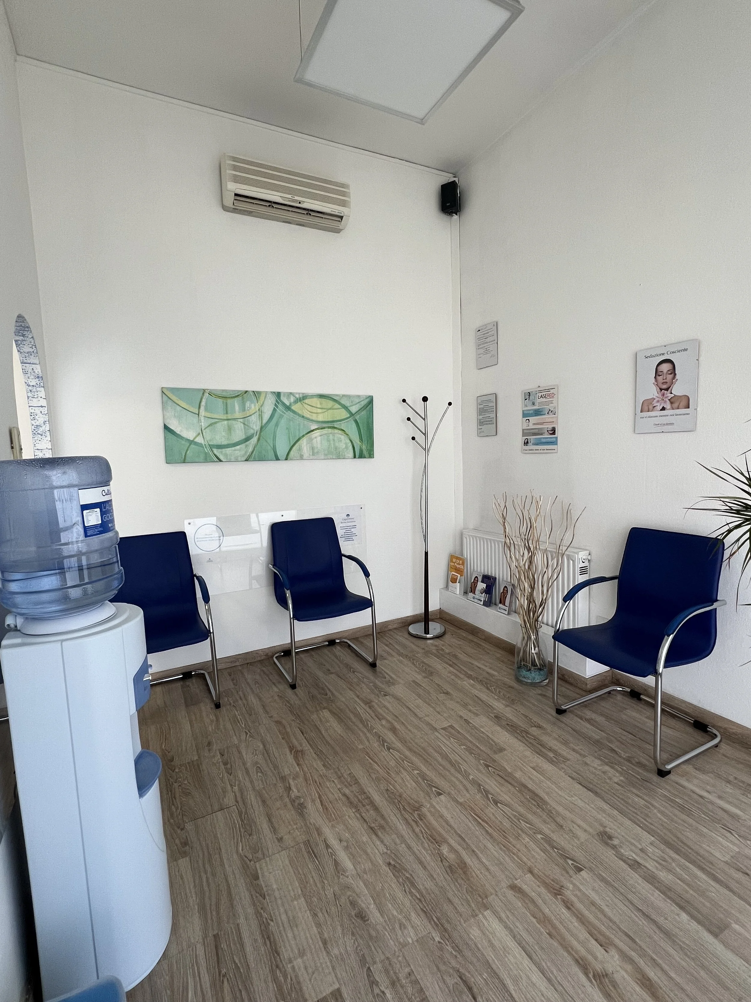 Sala d'attesa con tre sedie blu, stand con brochure, vaso di fiori secchi e aria condizionata.