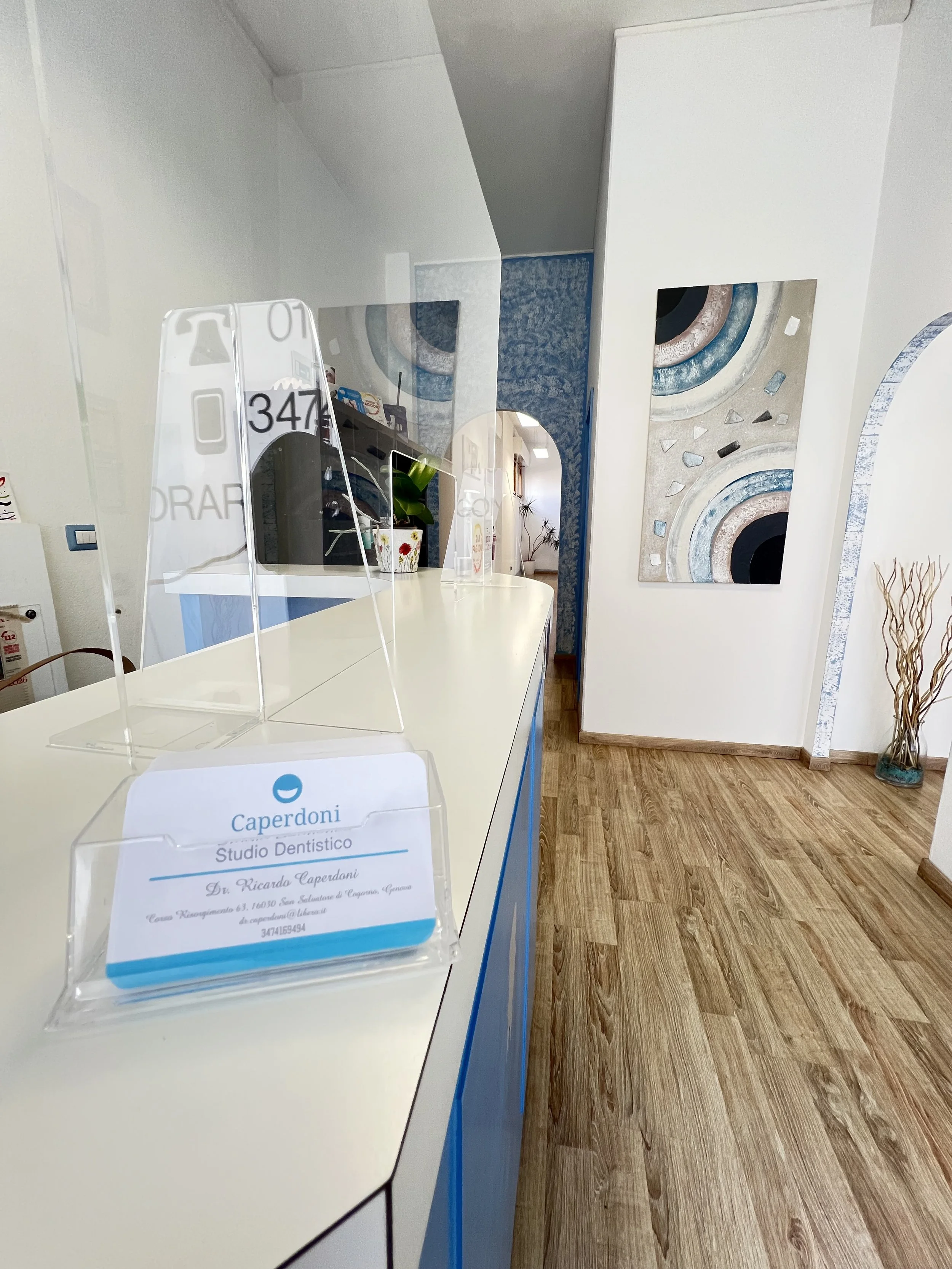 Reception area of a dental studio with a white counter, a transparent protective barrier, and a business card holder. The room has wooden flooring, colorful abstract wall art, and decorative plants.