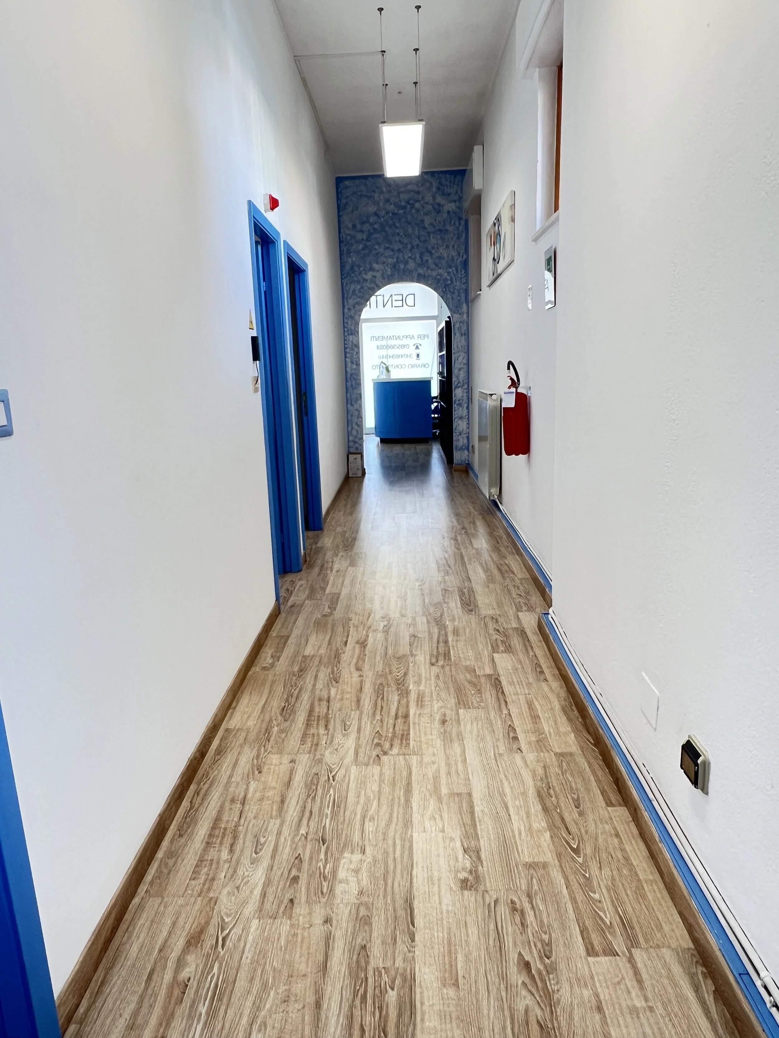 In questa immagine si vede un corridoio con pareti bianche, pavimento in legno e porte blu, con un'illuminazione centrale e un arco con scritte sulla vetrina alla fine del corridoio.