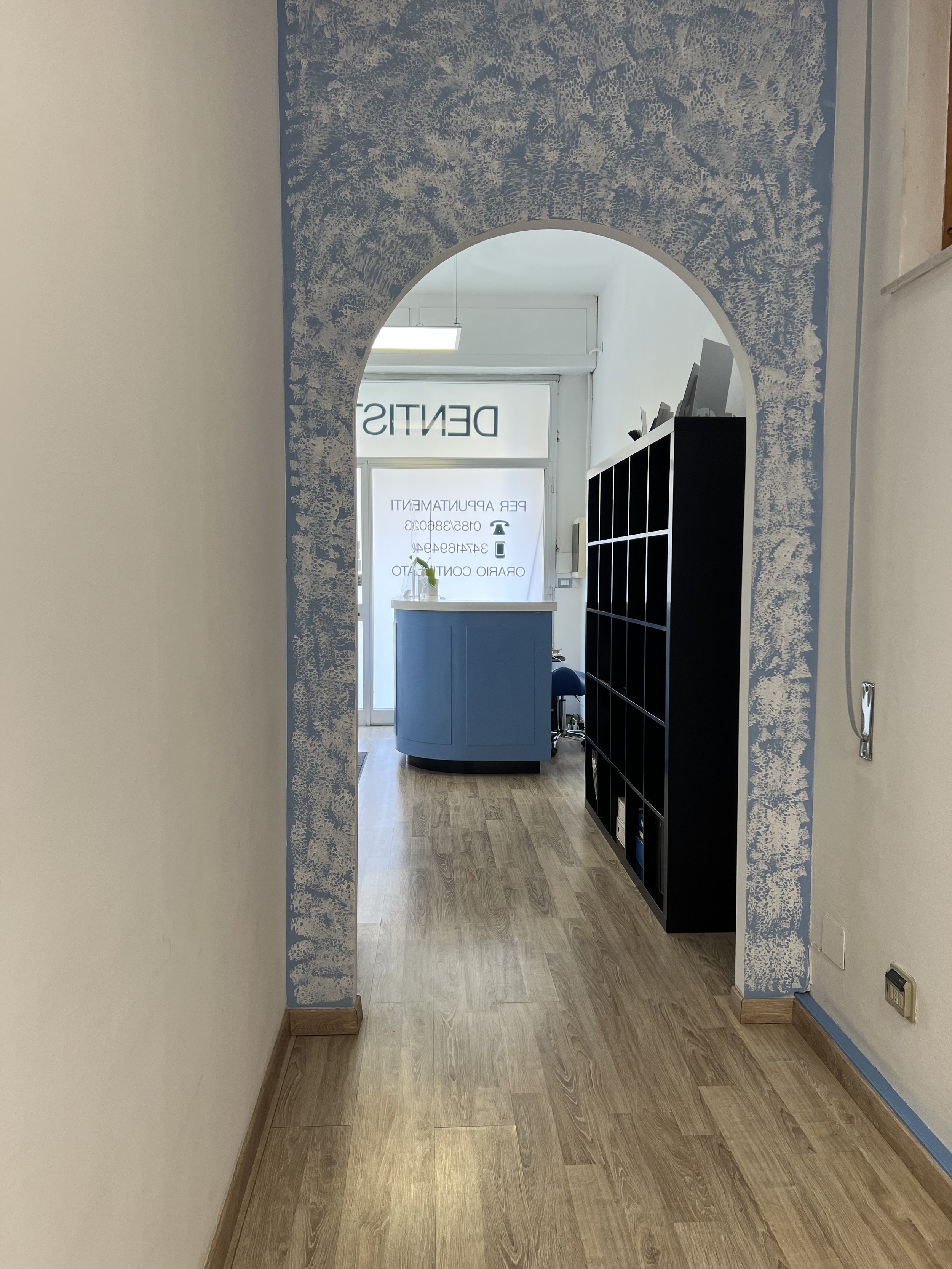 Ingresso di un ufficio con porta in vetro, scaffale nero e reception blu, vista attraverso un arco