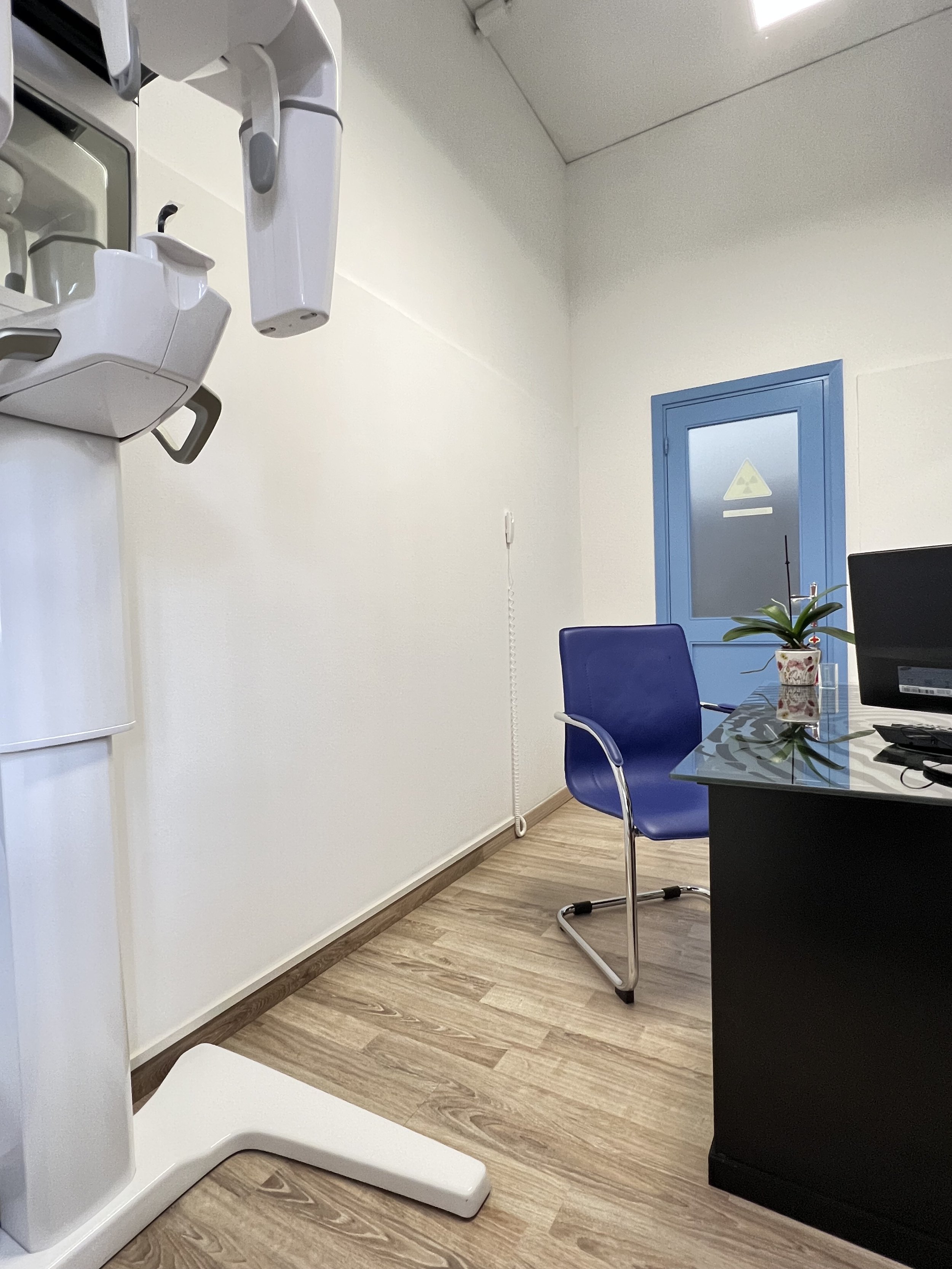 Stanza di uno studio dentistico con apparecchiature mediche, una sedia blu, un tavolo con un computer e una pianta sul tavolo, una finestra con simbolo di radiologia.