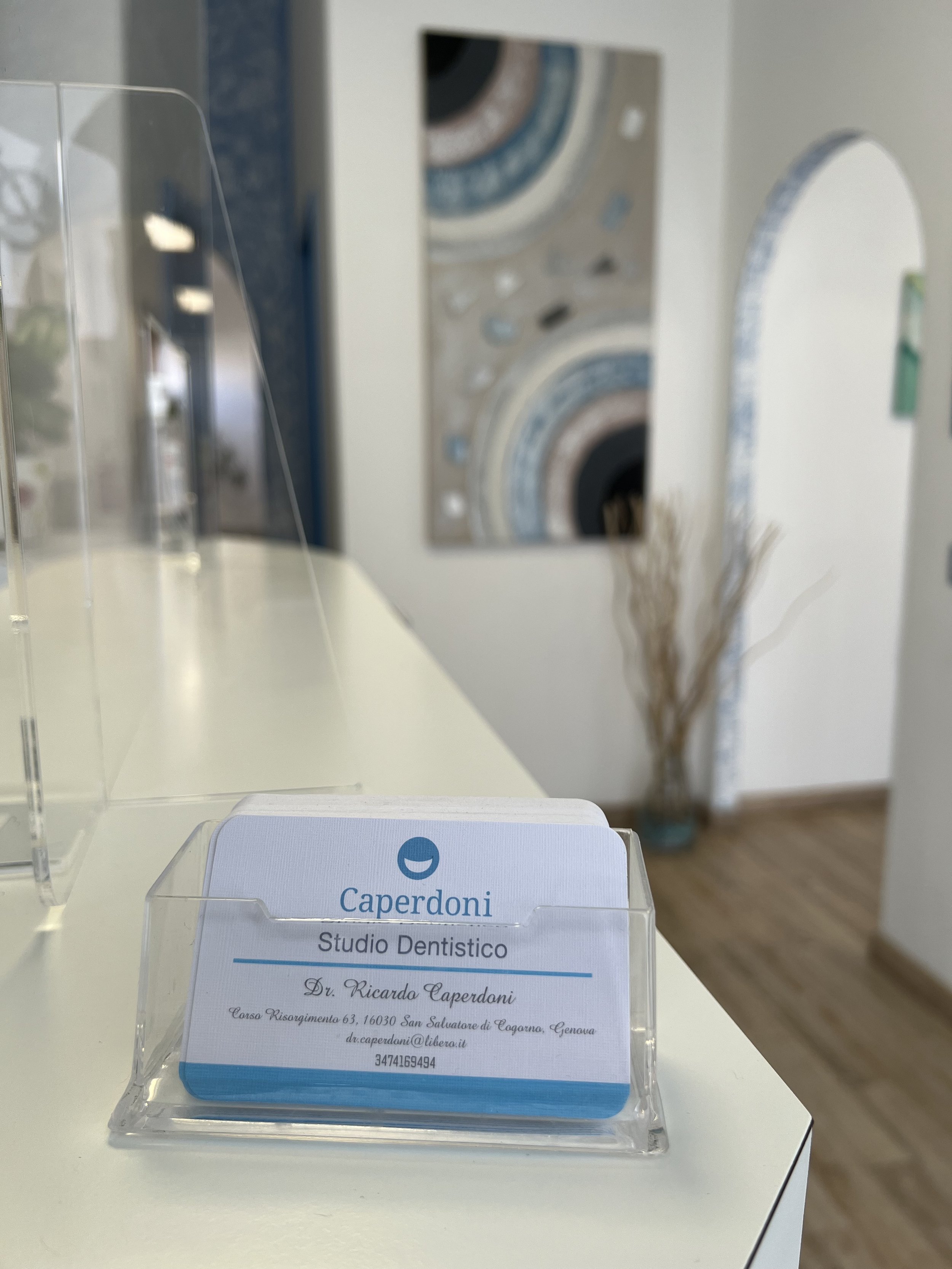 Biglietto da visita del dentista Dr. Ricardo Caperdoni, Studio Dentistico, con indirizzo e contatti, posizionato su un banco bianco in uno studio dentistico con decorazioni astratte sullo sfondo.