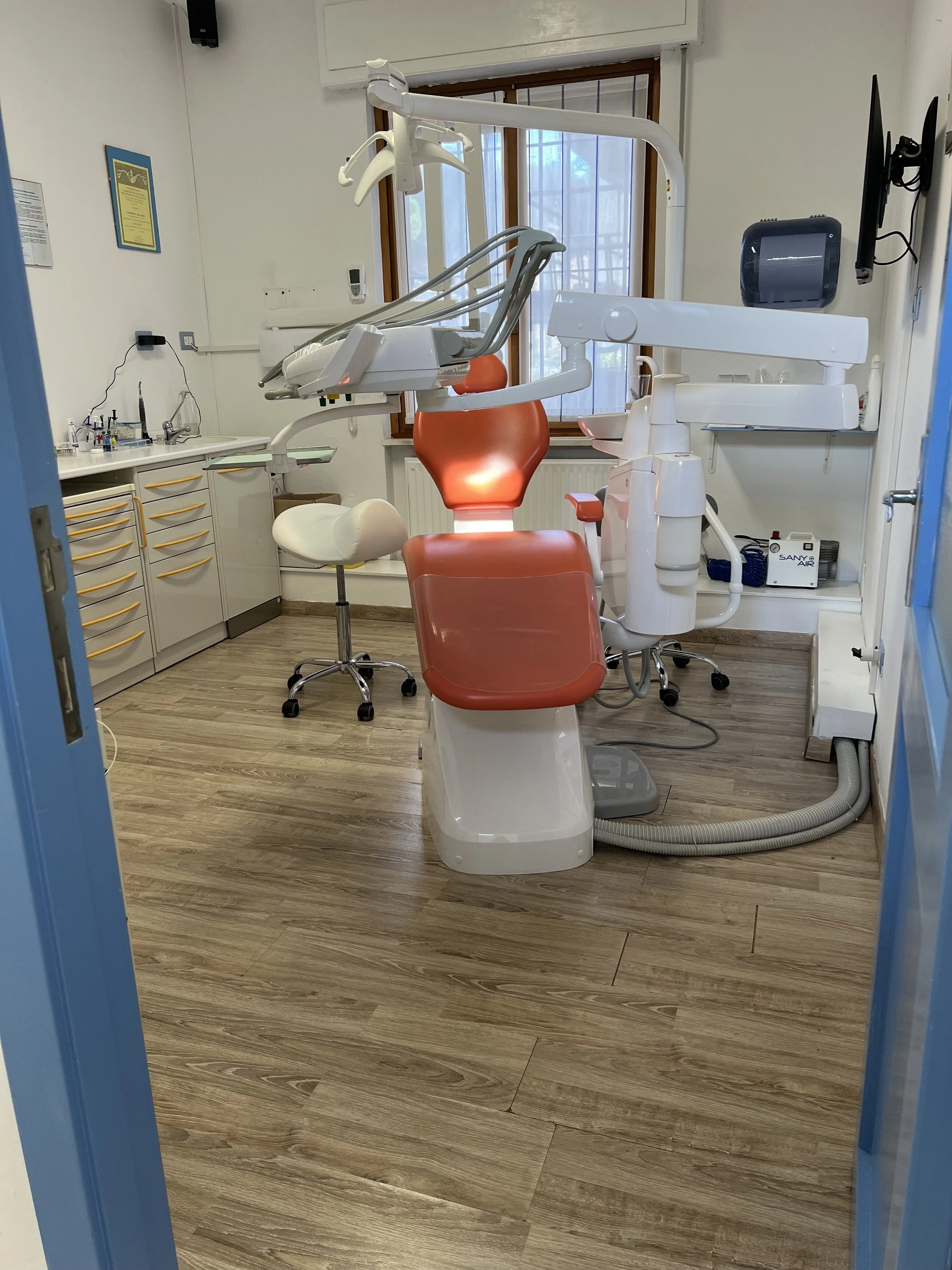 Stanza di studio dentistico con poltrona odontoiatrica rossa, strumenti dentali e attrezzature costantemente utilizzate, pareti bianche, finestra con tapparella, mobile con cassetti gialli, sedia da ufficio bianca, monitor a parete, e attrezzature mediche e di precisione.