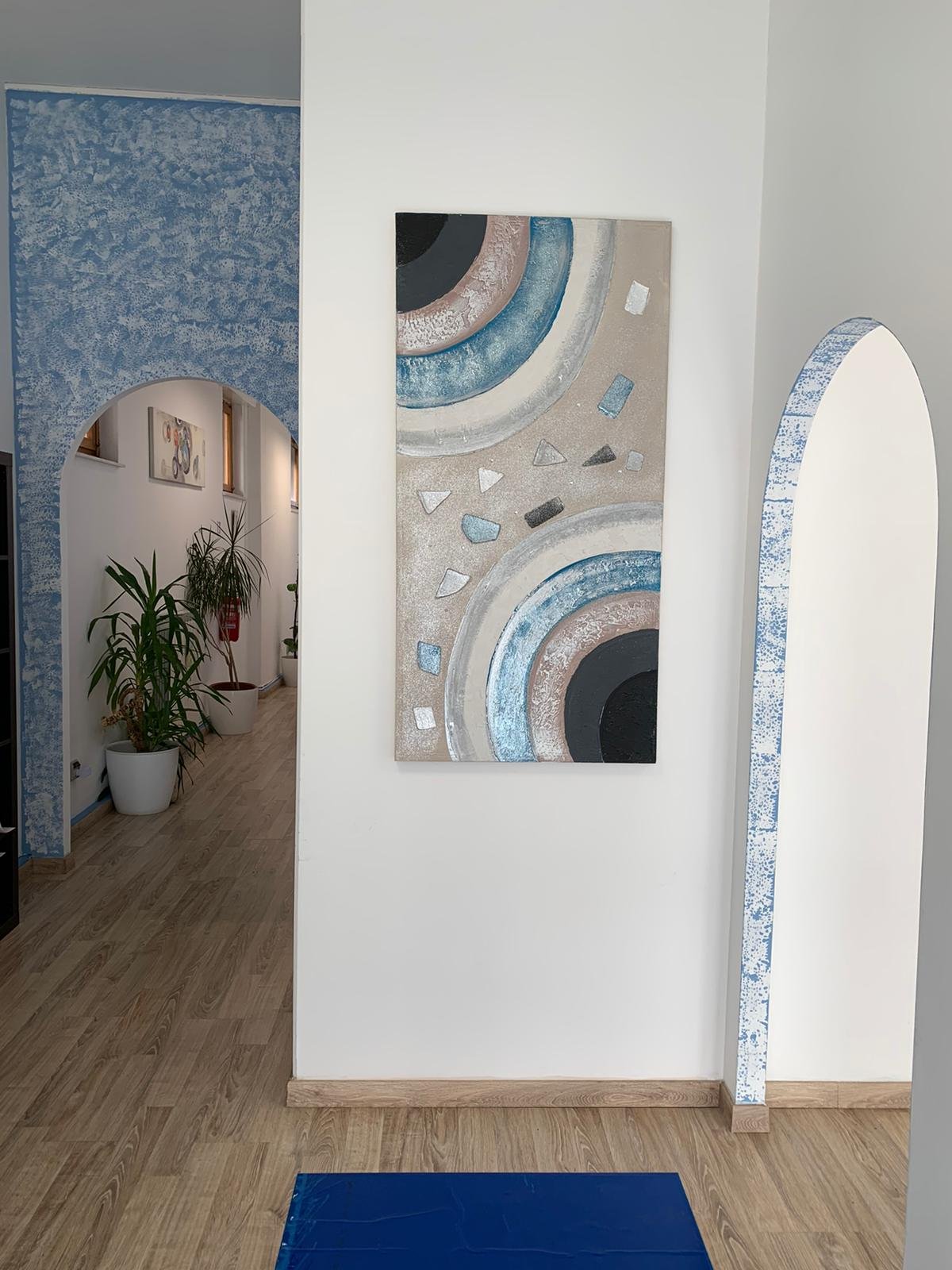 Interno di un edificio con parete bianca, decorata con un quadro astratto con cerchi concentrici blu, beige, nero e bianco. Sul lato destro, un arco decorato con mosaico blu e bianco. Sul lato sinistro, un corridoio con piante in vasi bianchi, pareti con quadri e pavimento in legno.