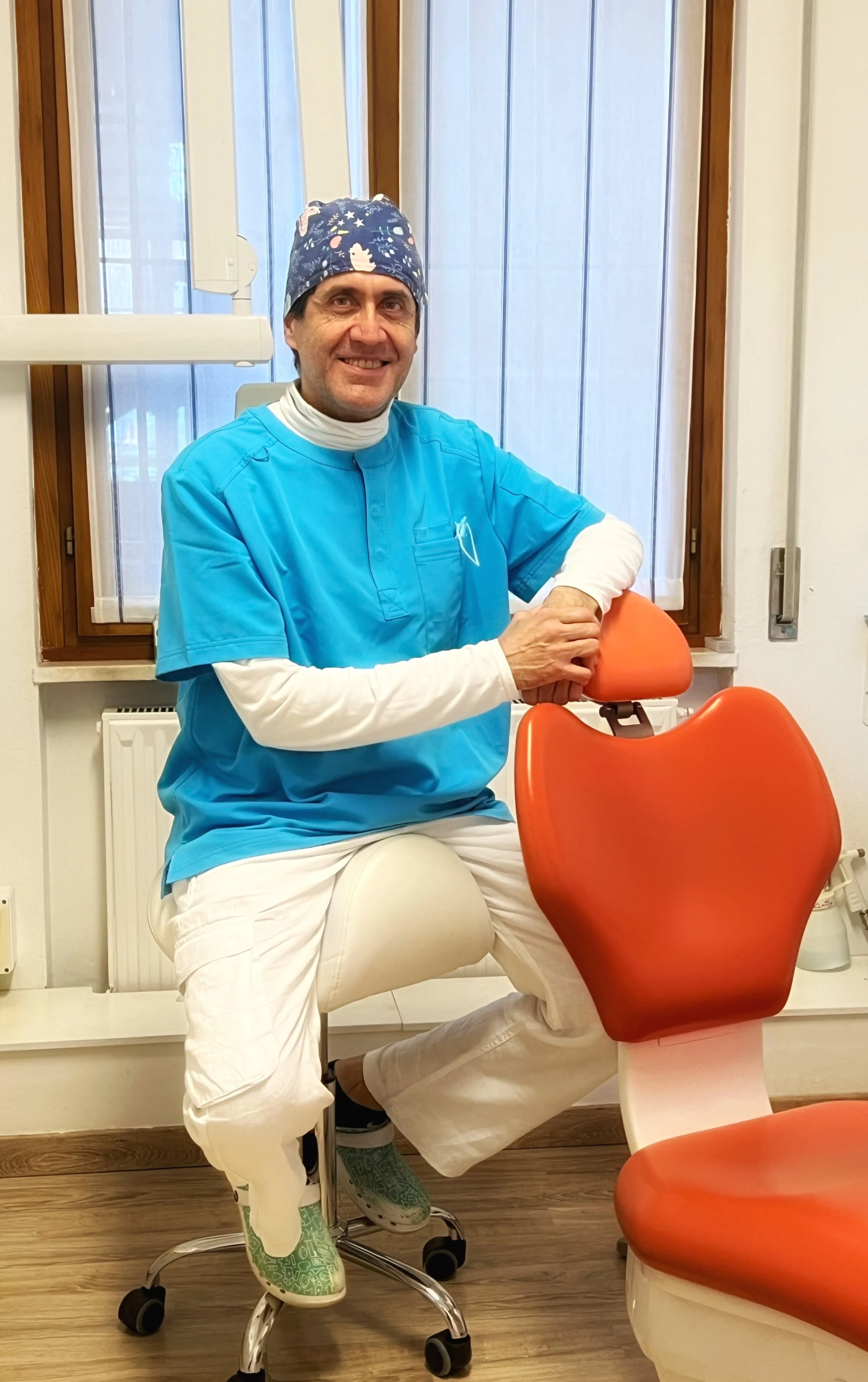 Un medico in divisa blu con un cappello colorato, seduto su una sedia da ufficio in una stanza con finestre con tende, sorridente, vicino a una sedia con sedile arancione.