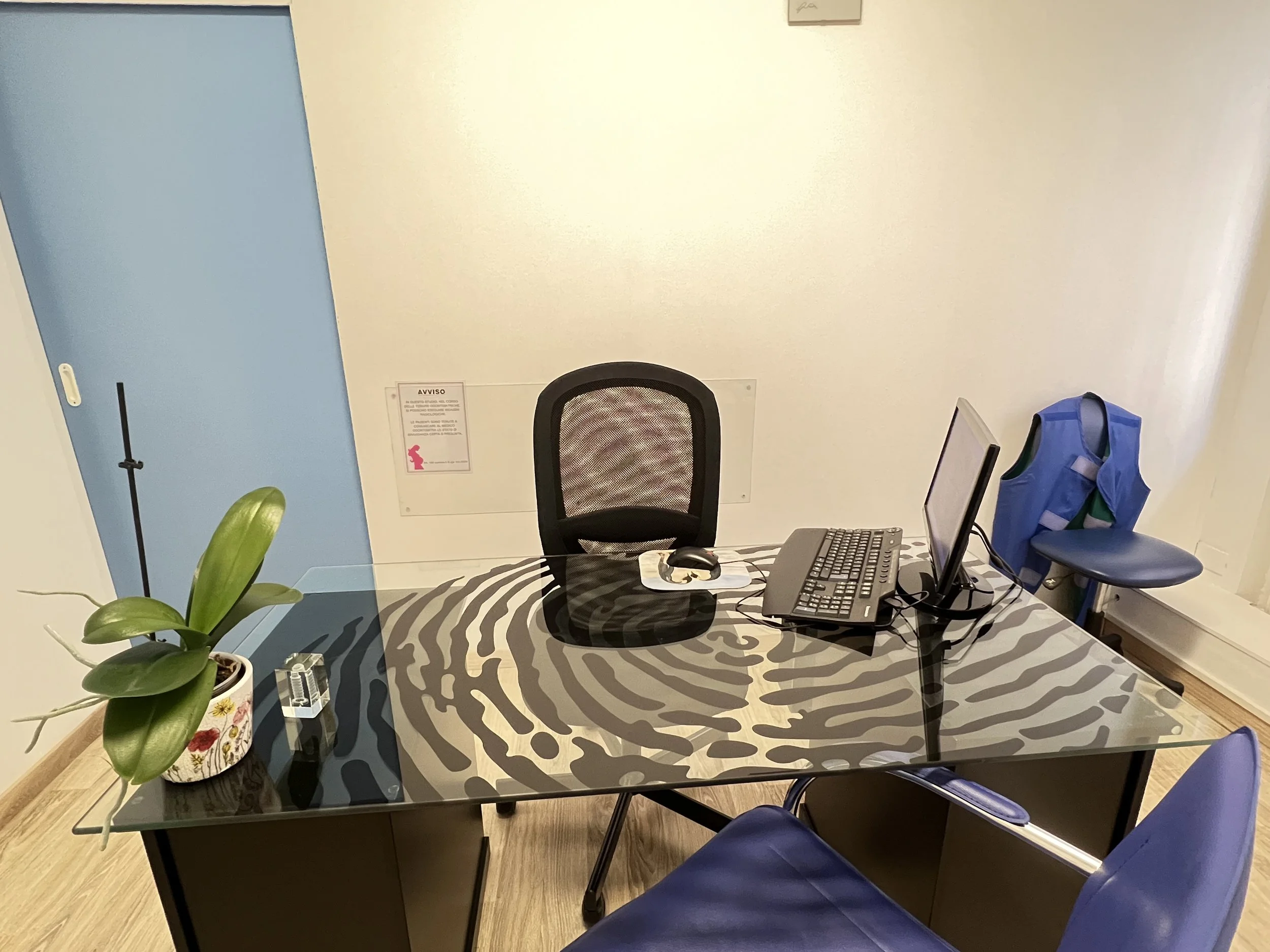 Una scrivania con una sedia, un computer e una pianta, in un ufficio moderno con pareti bianche e una porta blu.