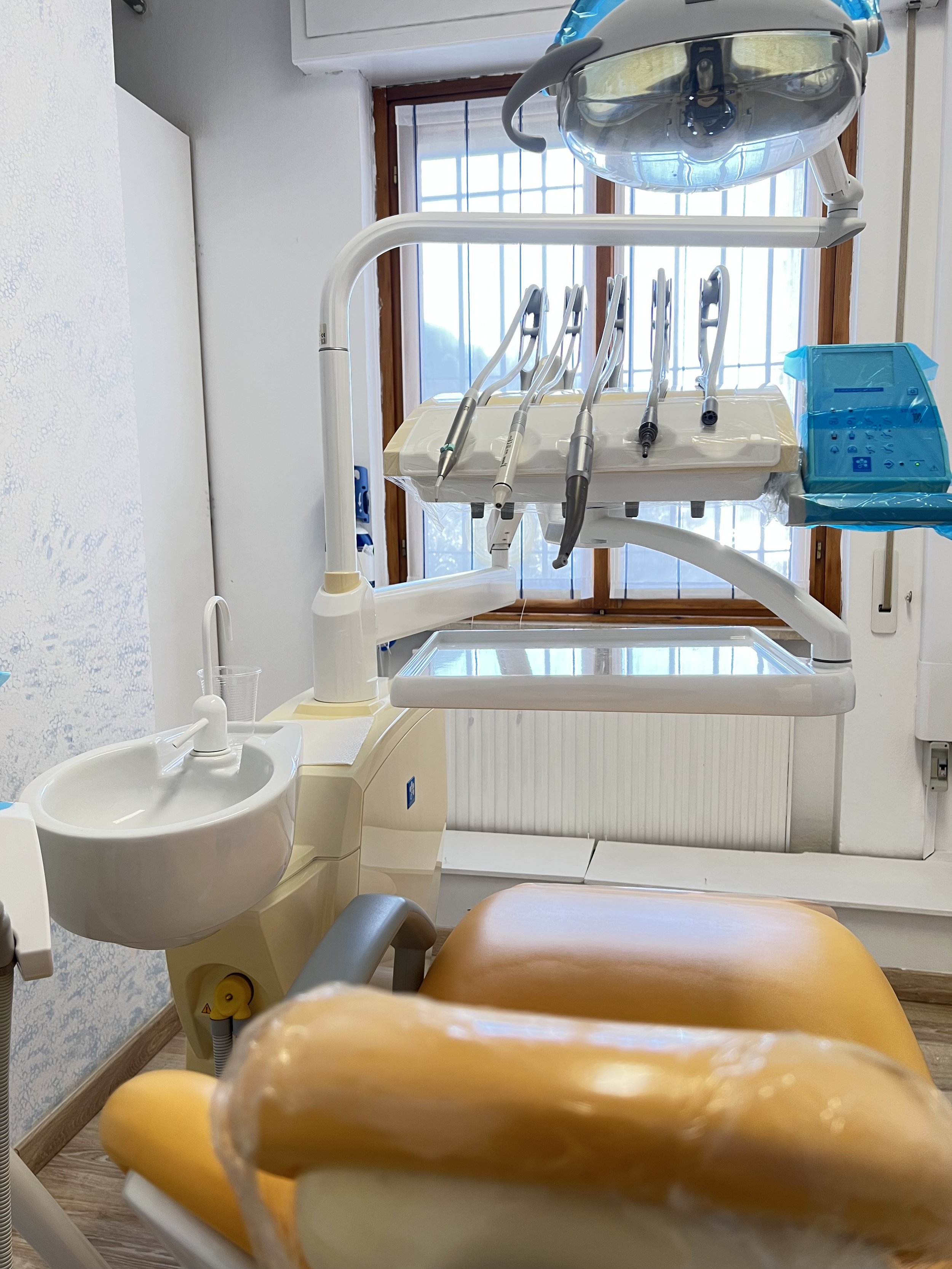 Sala di uno studio dentistico con le attrezzature. In primo piano, una poltrona obliqua di colore arancione, dietro la quale si vede un'unità dentale con strumenti e un'area di lavoro. Sullo sfondo, una finestra con grate in metallo.
