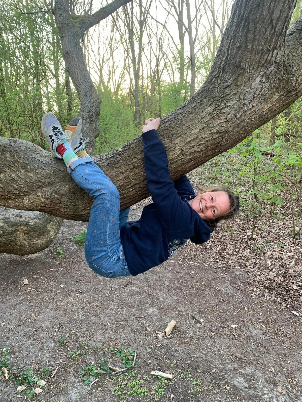 Vrouw hangt ondersteboven aan een boomtak in het bos, lachend en gekleed in een donkerblauwe jas en spijkerbroek.