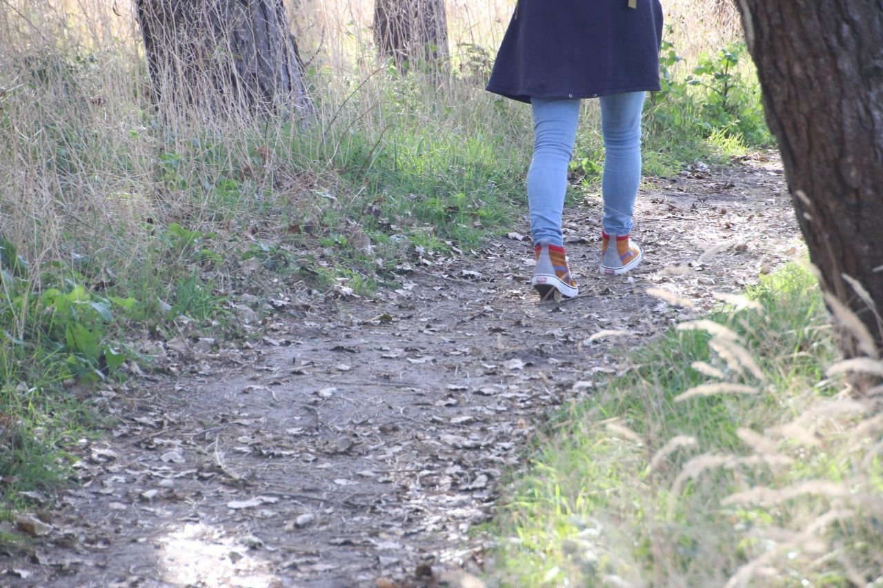 Persoon wandelt op een bospad, gefocust op de achterzijde met blauwe jeans, een zwart jas en sneakers.