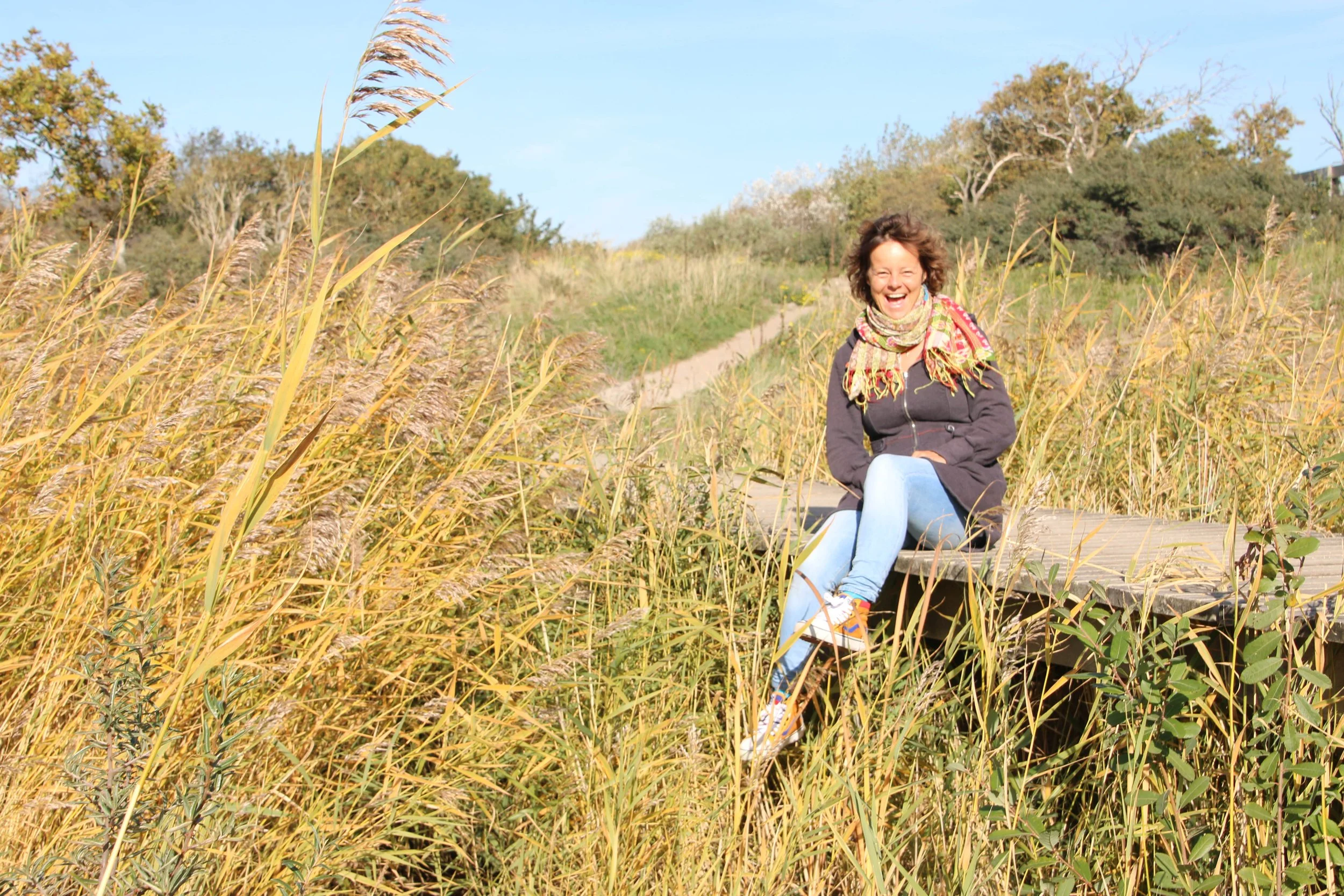 Vrolijke vrouw zittend op een houten brug in een veld met hoge, geelbruine grassen, onder een heldere blauwe hemel.