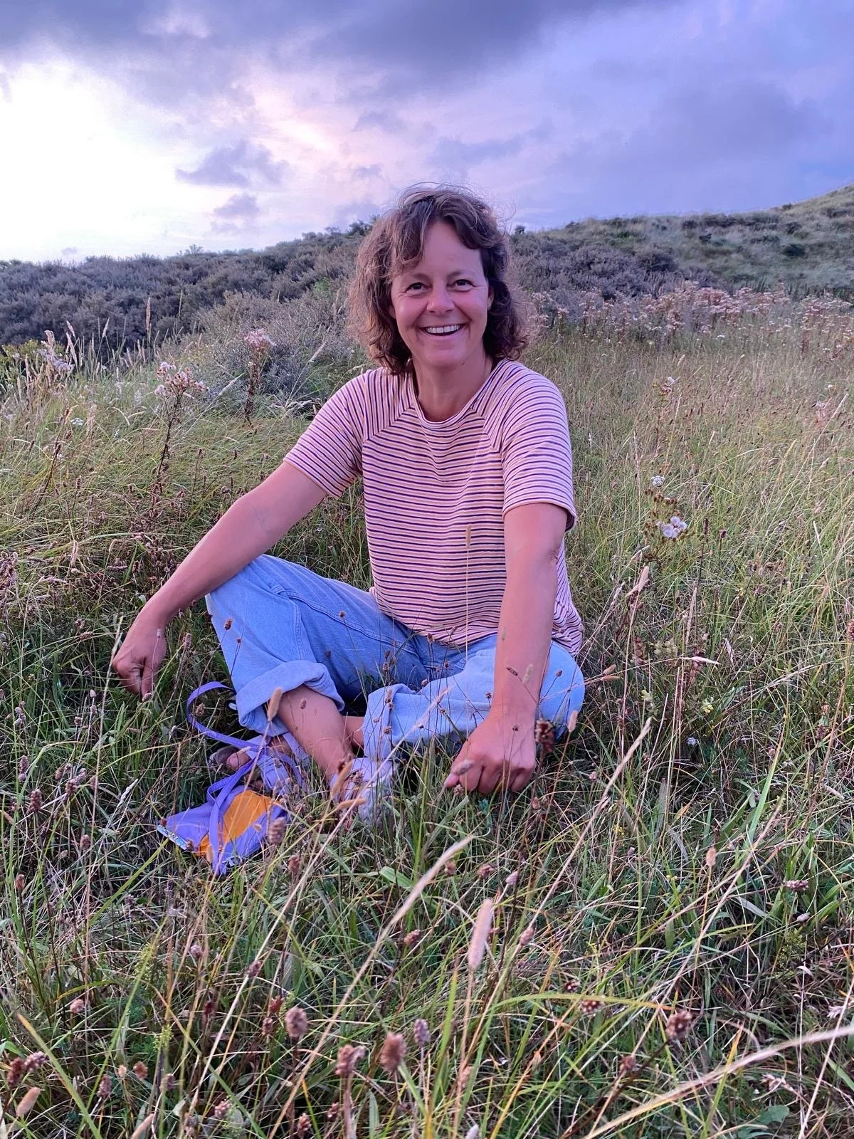 Een glimlachende vrouw zit op een grasveld in de natuur, omgeven door bloemen en lage struiken, met een heuvel en een bewolkte hemel op de achtergrond.