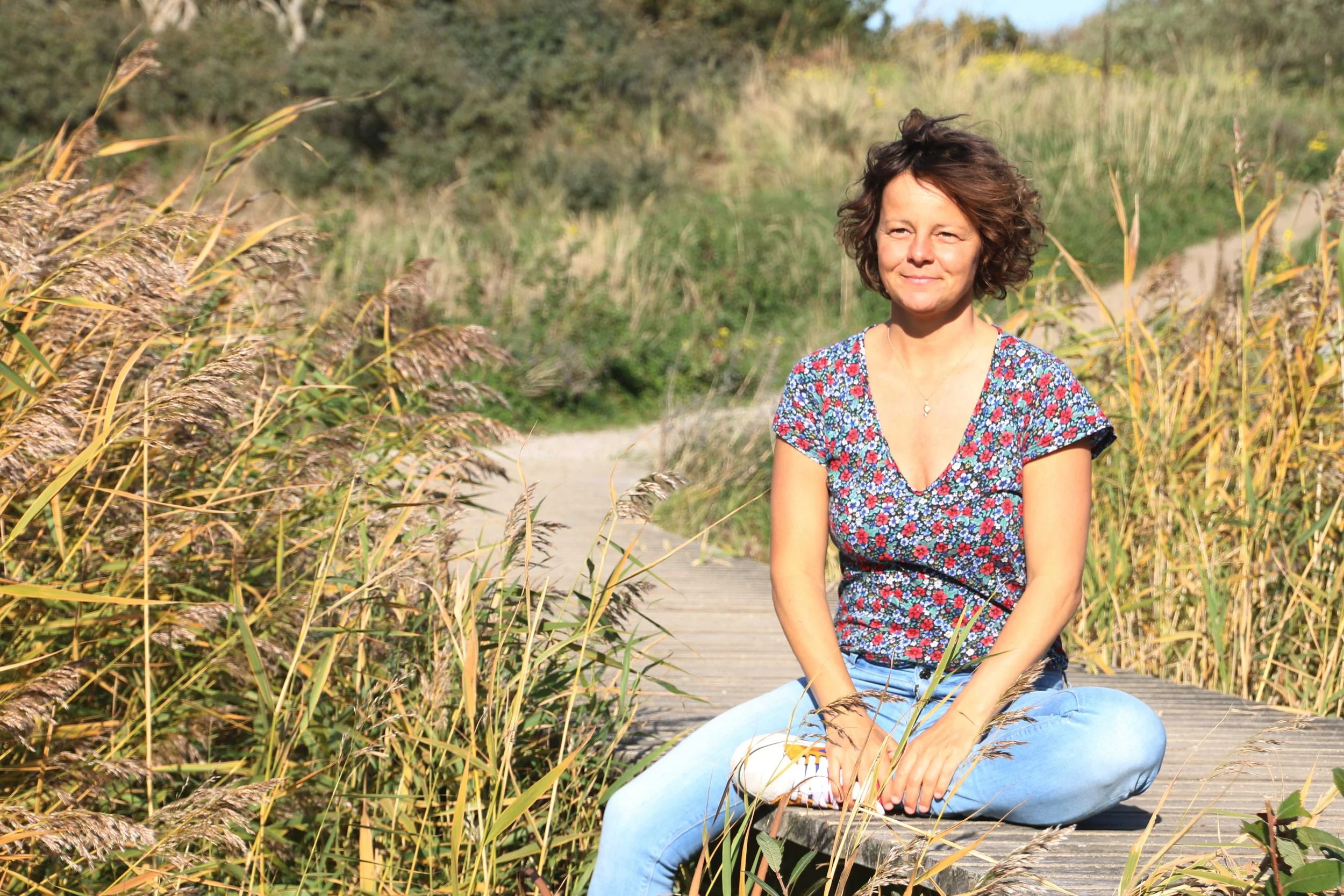 Vrouw zit gekruist op een houten pad in een natuurlijke omgeving met gras en struiken, mogelijk in de herfstperiode.