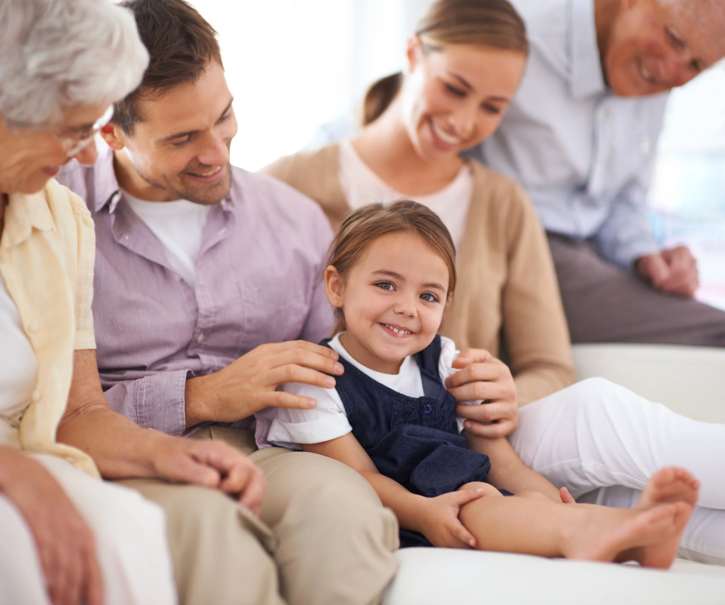 A family of six people, including three generations, sitting together on a white couch, smiling and enjoying each other's company.
