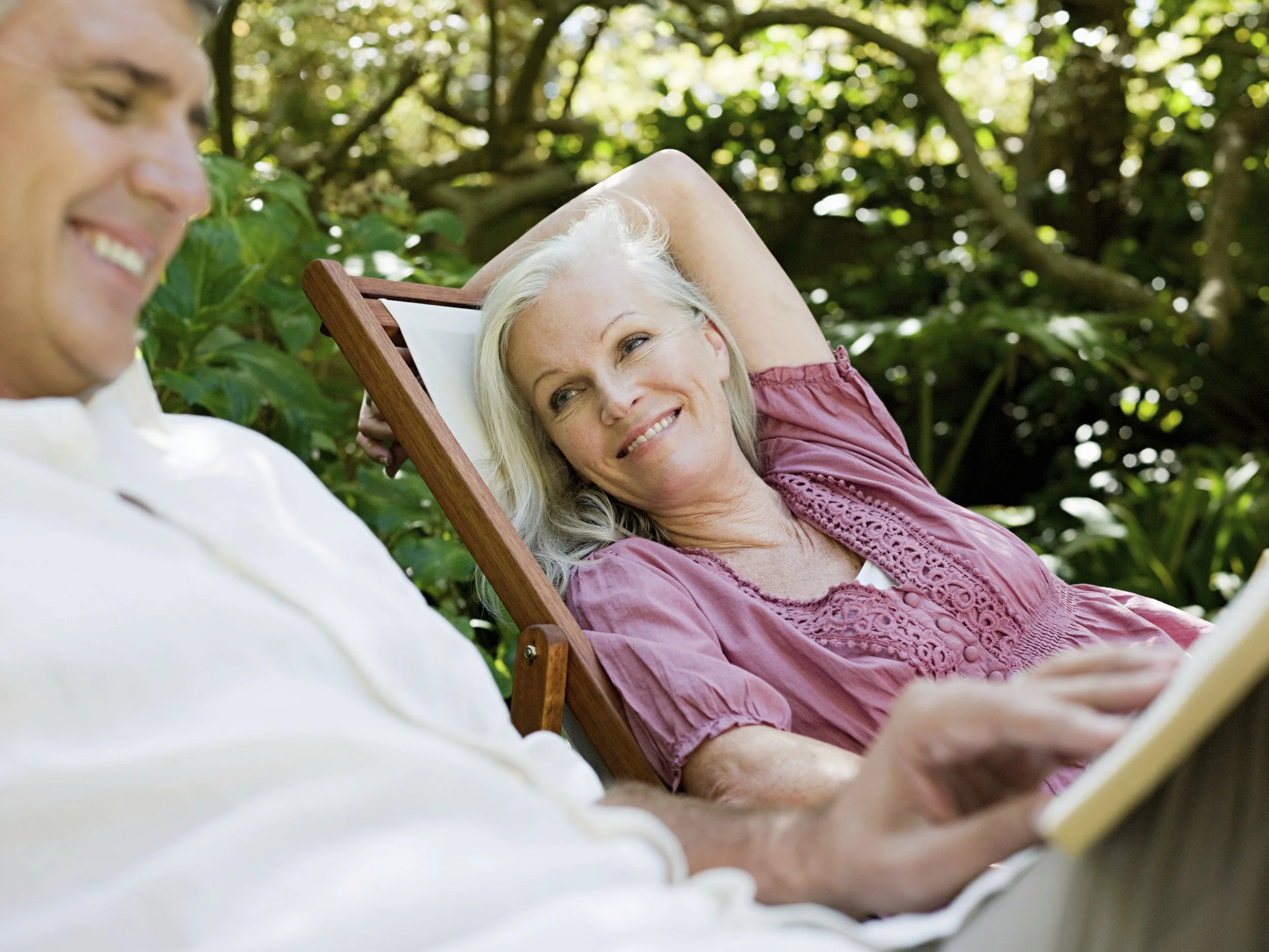 A woman with gray hair in a pink blouse relaxing on a wooden lounge chair outdoors, smiling, while a man, partially visible, interacts with her.
