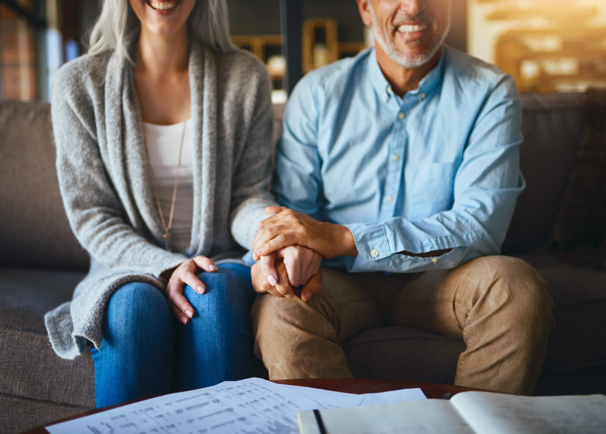 A close-up of a couple sitting on a sofa, holding hands, with papers and a notebook on the table in front of them.