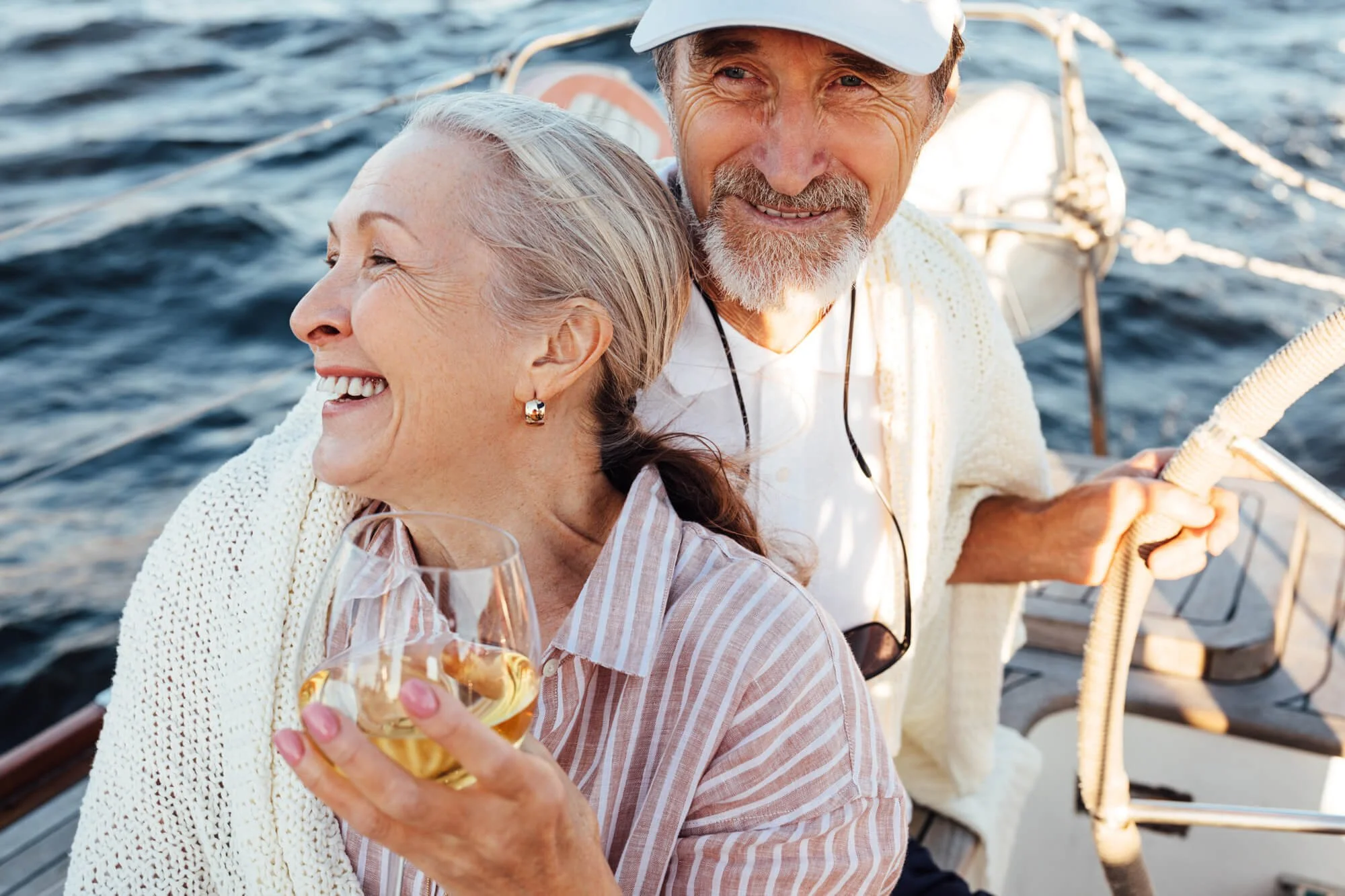Senior couple enjoying a boat ride, smiling and holding a glass of white wine on a sunny day.