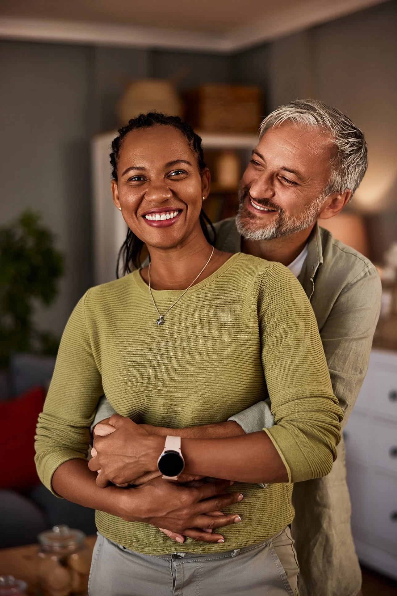 A smiling woman in a yellow-green sweater and a man with gray hair and beard hugging her from behind in a cozy living room.