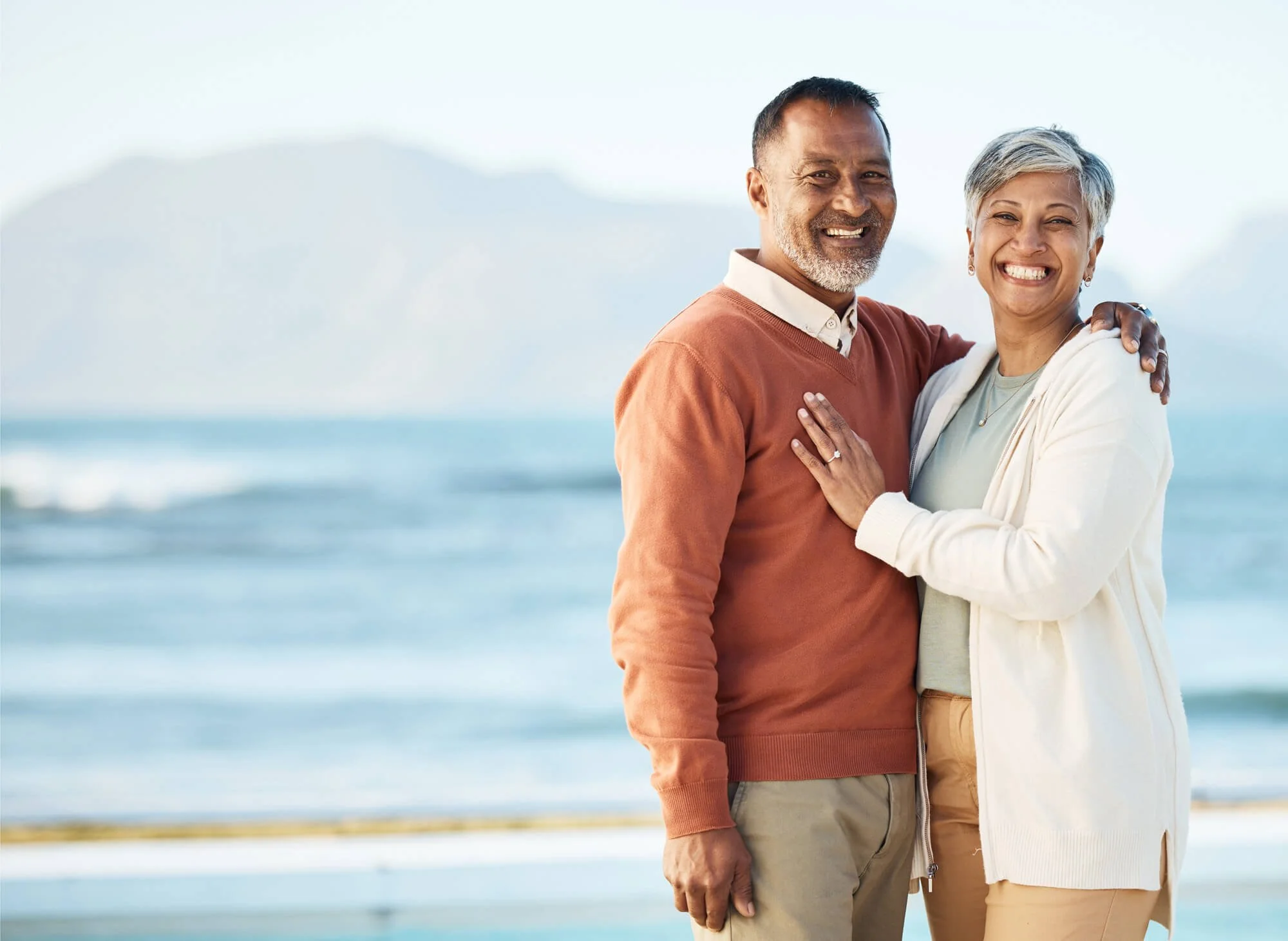 A happy older couple stands on the beach with ocean waves and a mountain in the background, smiling and embracing.