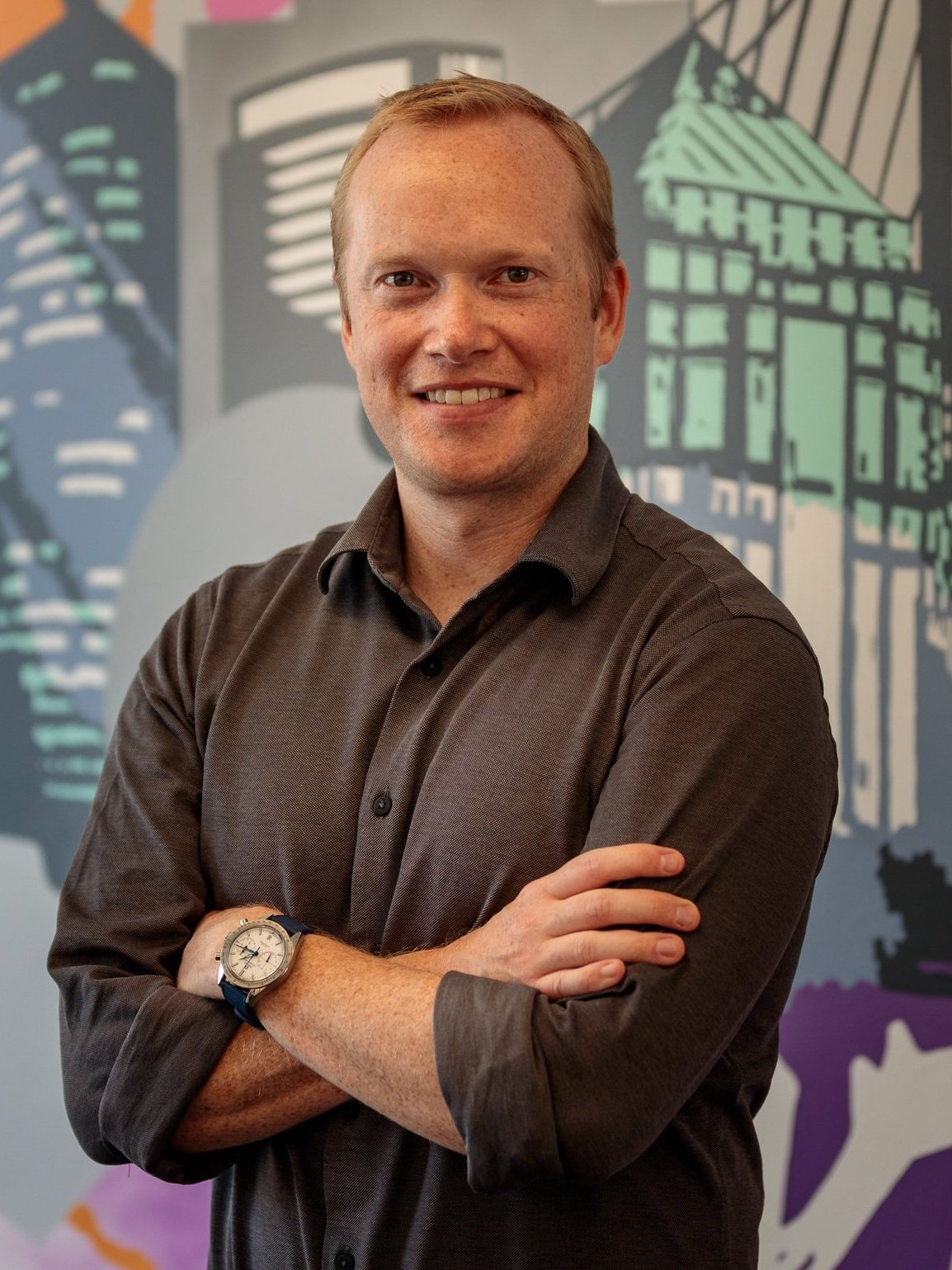 A young man with short blonde hair, freckles, and a big smile, standing with arms crossed in front of a colorful mural of city buildings and landmarks.