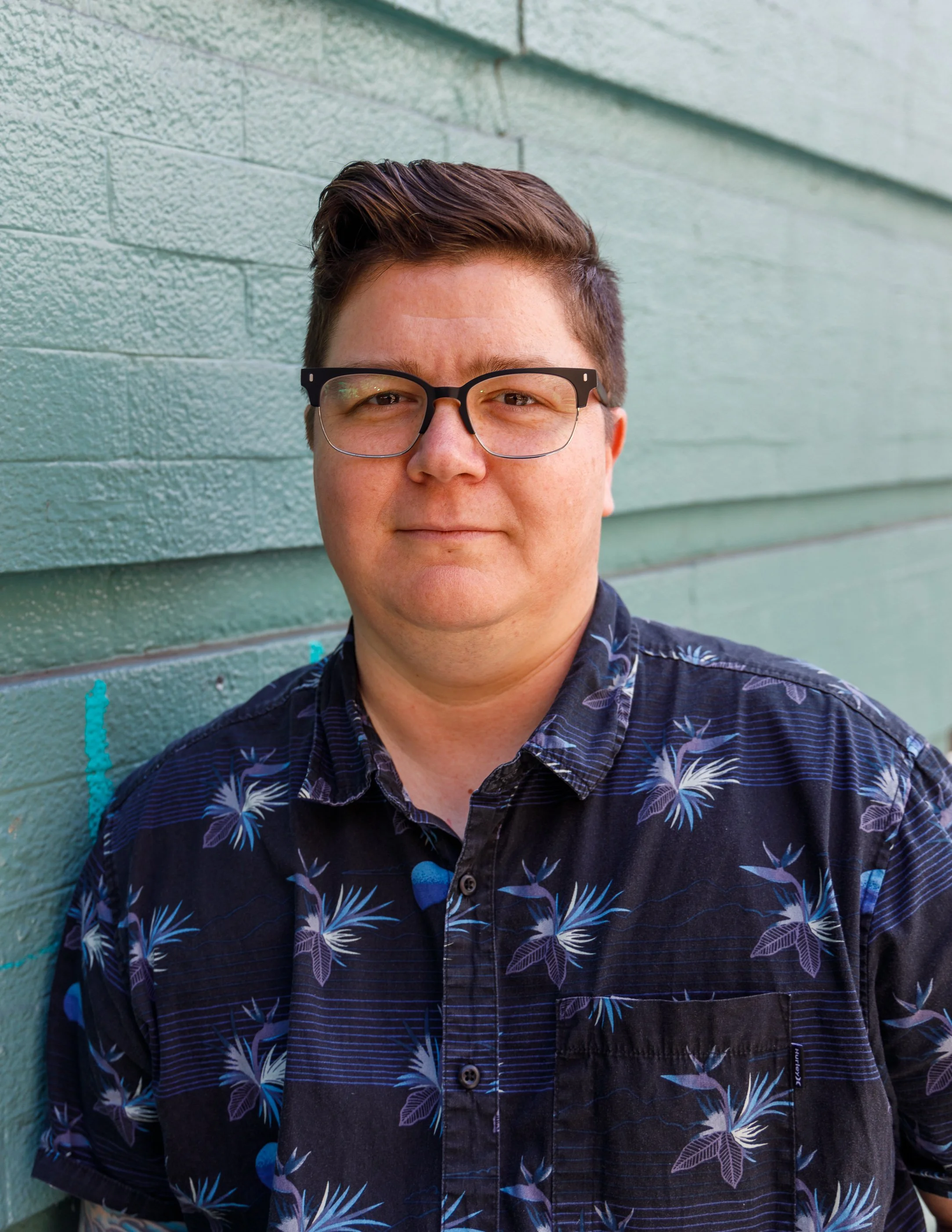 A man with short brown hair, wearing glasses, and a dark Hawaiian shirt with a floral pattern, standing against a green brick wall.