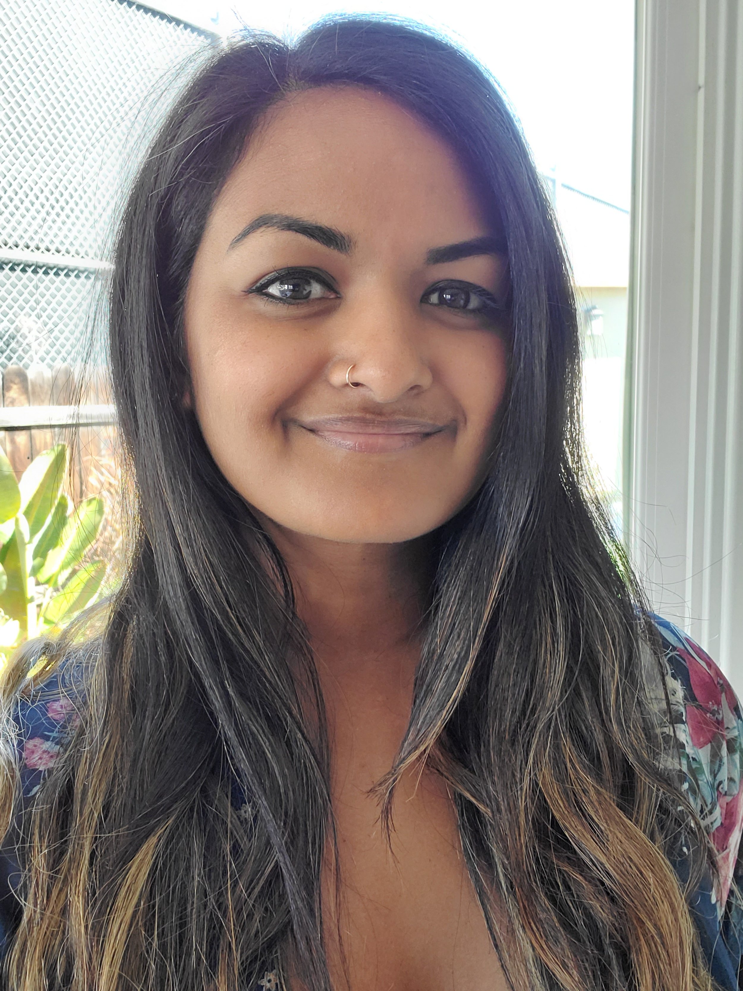 A young woman with long dark hair, light makeup, and a nose ring, smiling in front of a window with sunlight, plants, and a lattice fence outside.